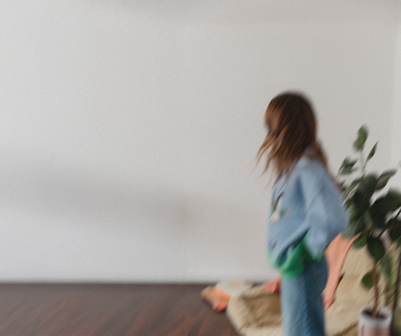 Emily Rose wearing a blue jacket and jeans, standing near a beige sofa with colorful pillows, a large potted plant, and a plain white wall in the background.