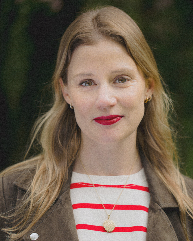 Katie Williams with long blonde hair wearing a brown jacket, a red and white striped shirt, and red lipstick, outdoors with a blurred green background.