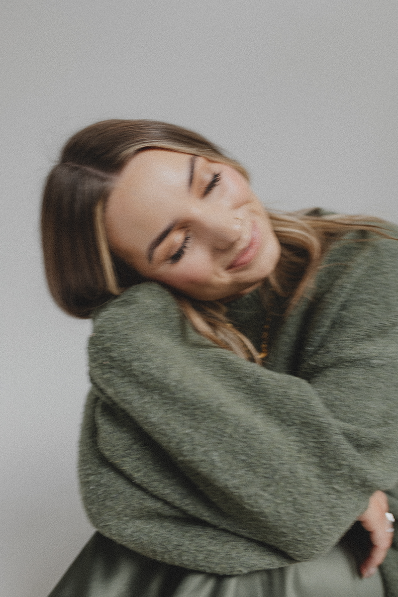 Emily Rose with light brown hair and closed eyes, smiling gently, wearing a loose green sweater, facing slightly to the right, with a neutral background.