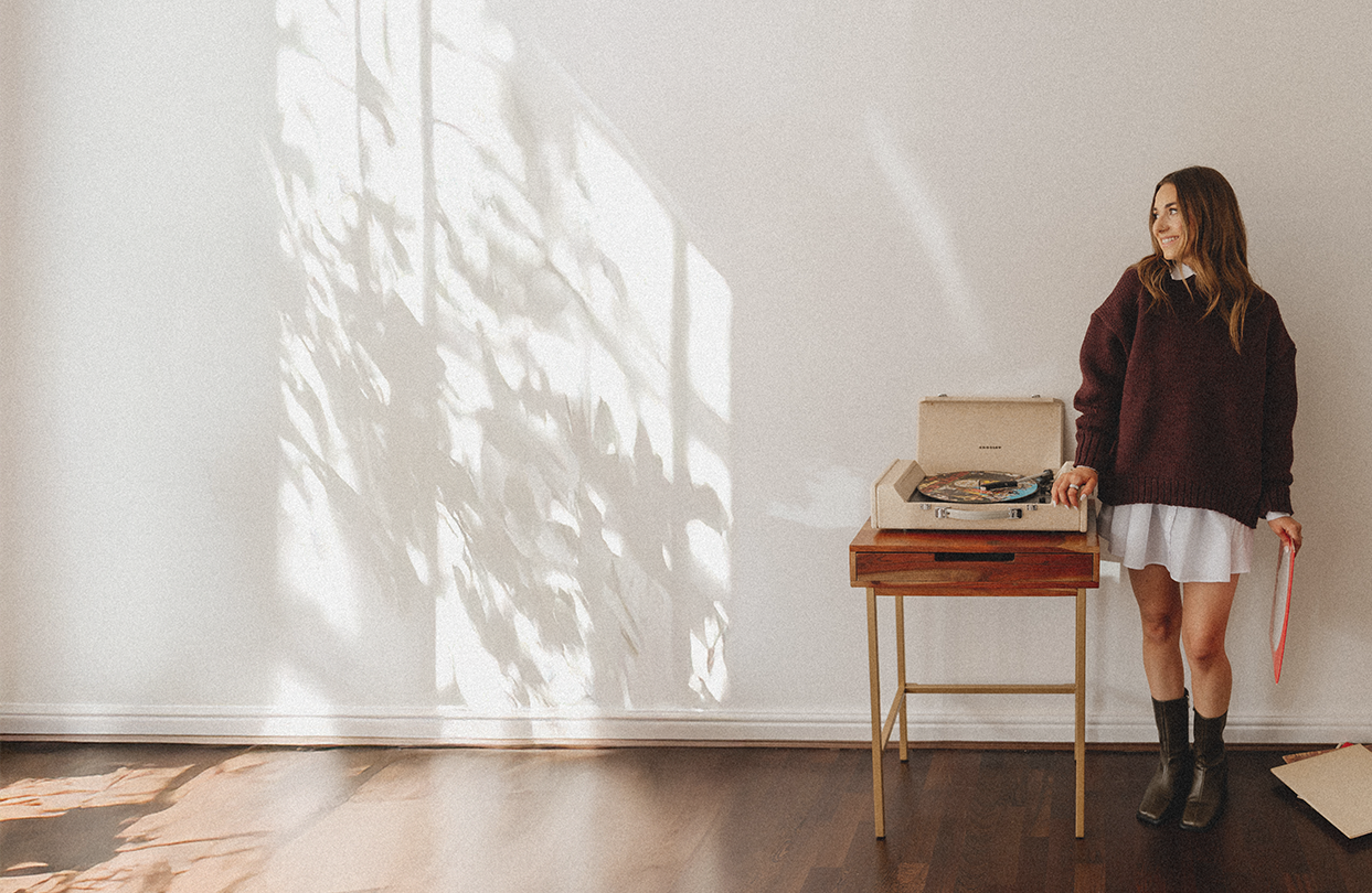 Emily Rose standing next to a vintage record player on a wooden table, holding a red record in a brightly lit room with shadow patterns on the wall.
