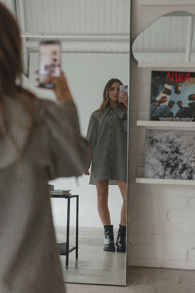 Emily Rose taking a mirror selfie in a room with white walls, wearing a checked dress and black ankle boots.