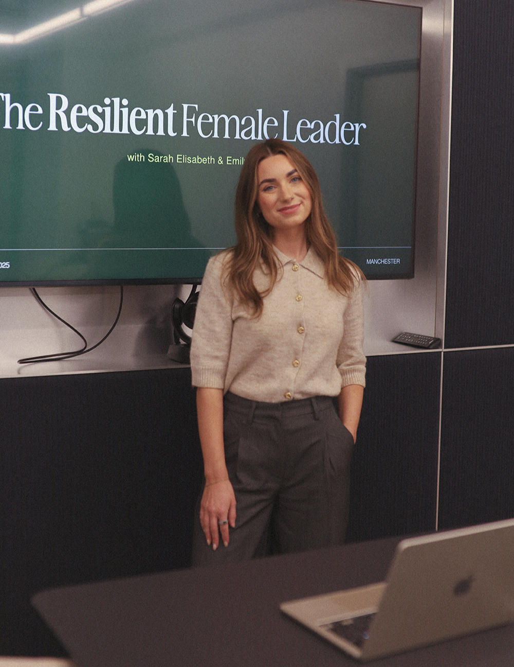 Emily Rose standing in front of a presentation screen that reads 'The Resilient Female Leader' with the names Sarah Elisabeth and Emily. She is wearing a beige button-up sweater and dark gray pants, smiling with her hands in her pockets.