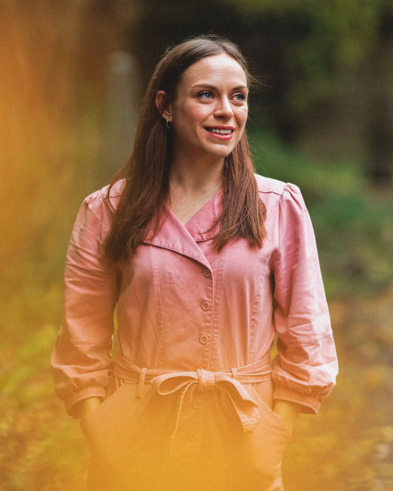 Lizzy Delaney with brown hair in a pink jacket outdoors, smiling, with green foliage in the background.