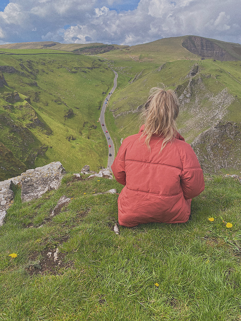 Emily Rose in a red jacket sitting on a grassy hill, overlooking a winding road through green hills and mountains under a partly cloudy sky.
