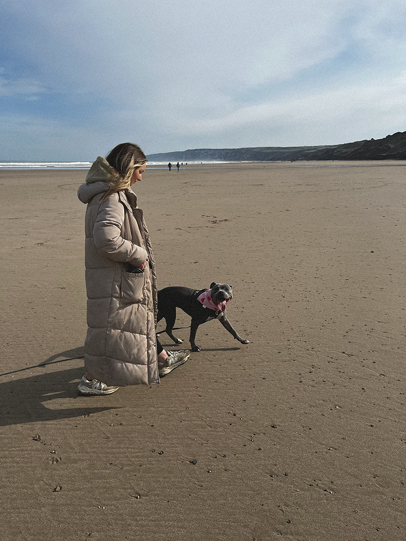 Emily Rose walking a black dog along a sandy beach, with the ocean and cliffs in the background.