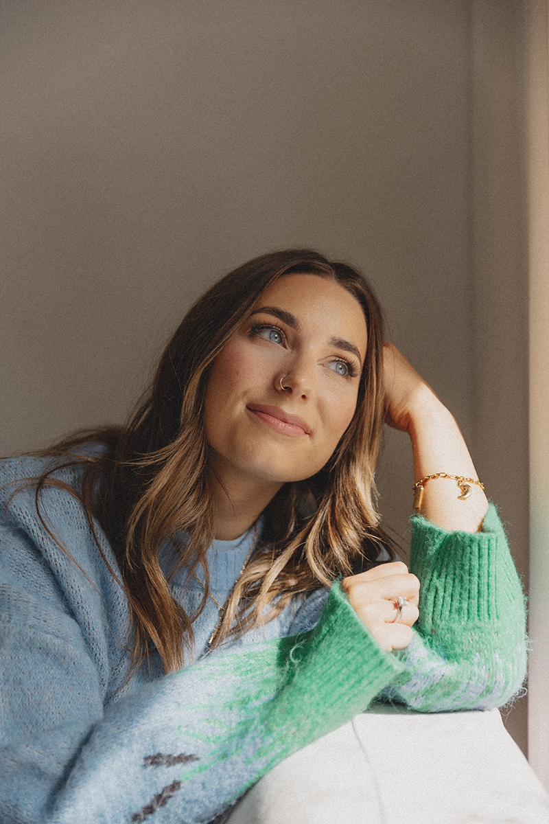 Emily Rose with long brown hair, blue eyes, and a nose ring, sitting by a window and looking outside with a gentle smile. She is wearing a blue sweater with green and brown patterns on the sleeves and a gold bracelet.