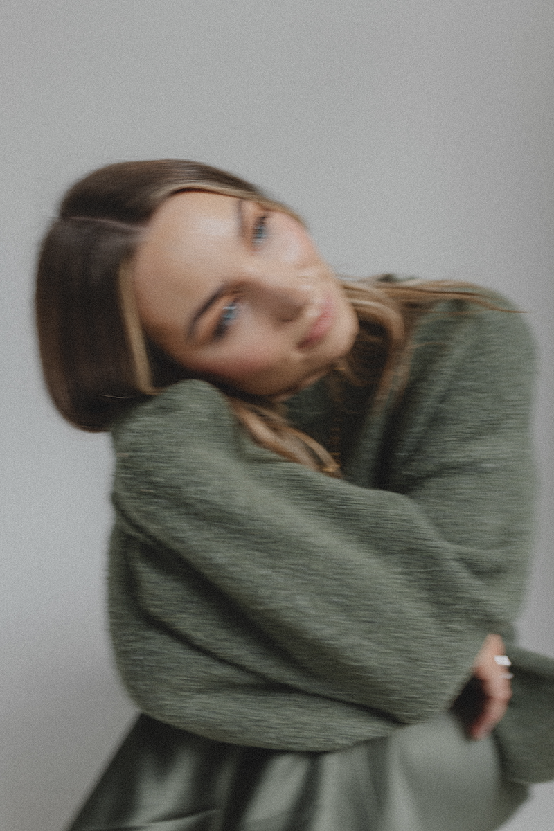 Emily Rose wearing a green sweater, is sitting with her head tilted and resting on her knee.