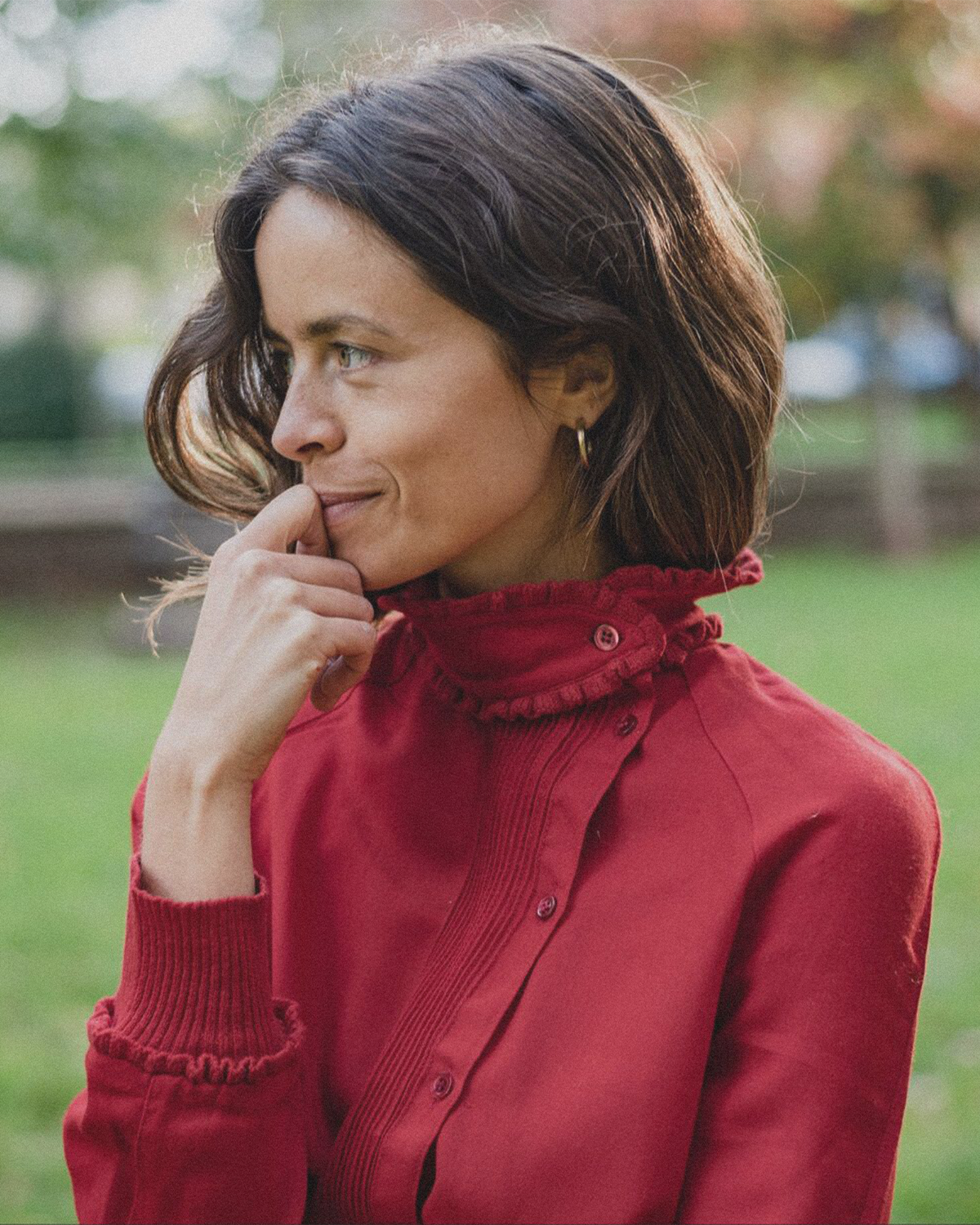 Ellen Mulcrone wearing a red jacket with a ruffled collar, looking to the left with a thoughtful expression and her hand near her mouth, outdoors with blurred green trees in the background.