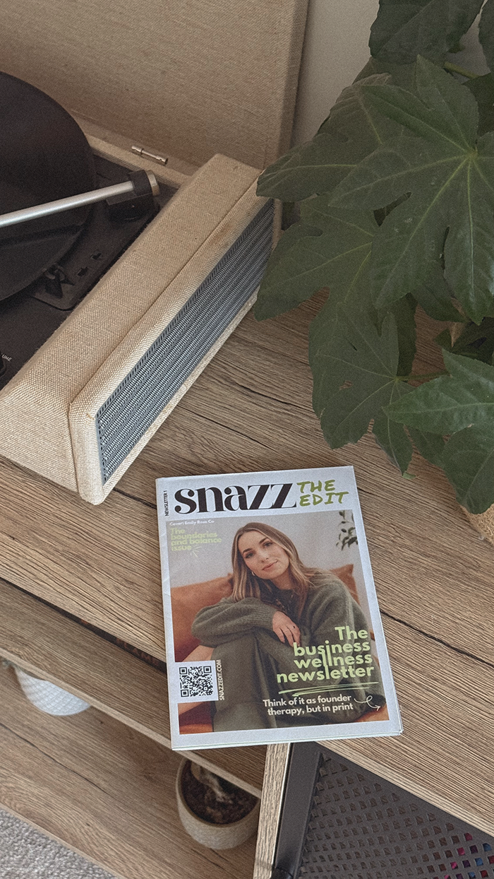 Snazz the edit magazine laying on a wooden surface next to a large green leafy plant, with part of a beige speaker and electronic device visible nearby.