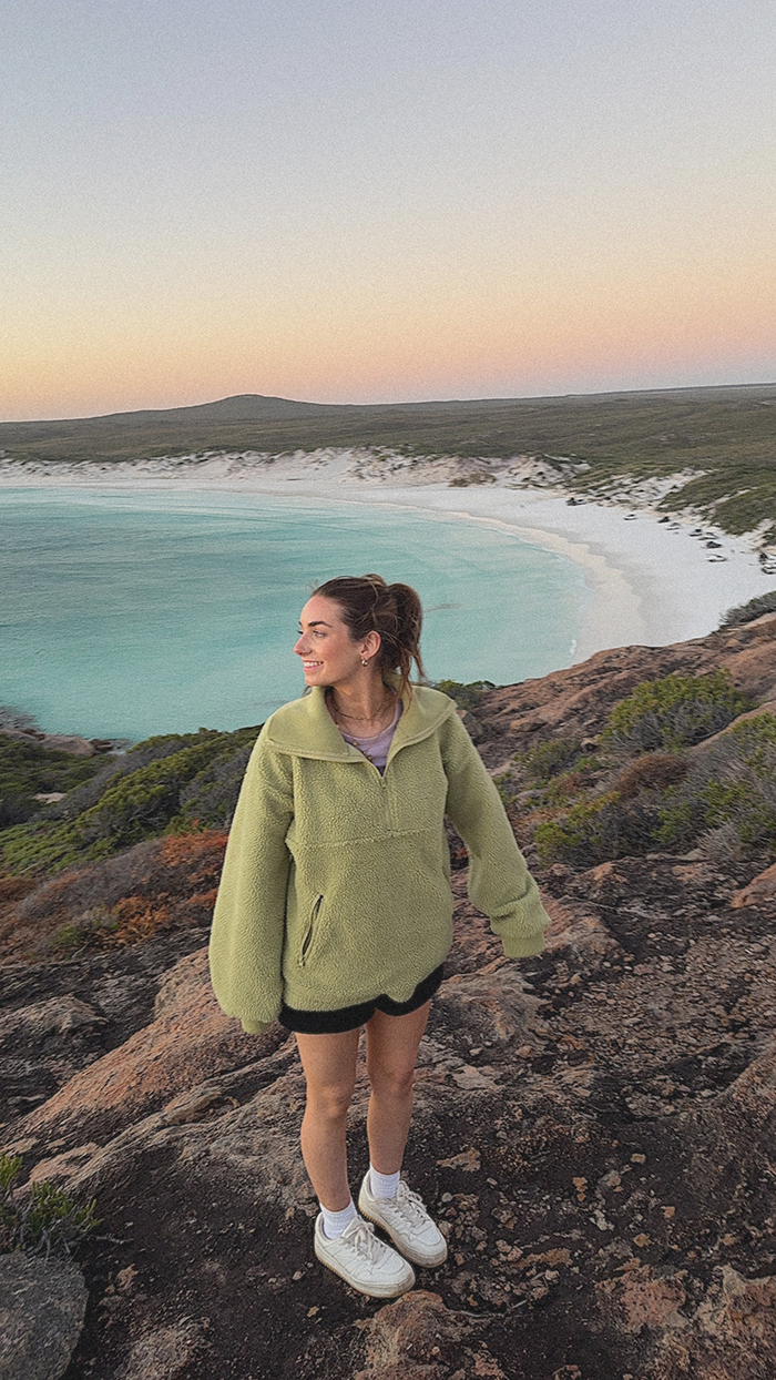 Emily Rose standing on rocky terrain near a beach with white sand and turquoise water during sunset, wearing a lime green fleece jacket, shorts, and sneakers, smiling while looking to the side.