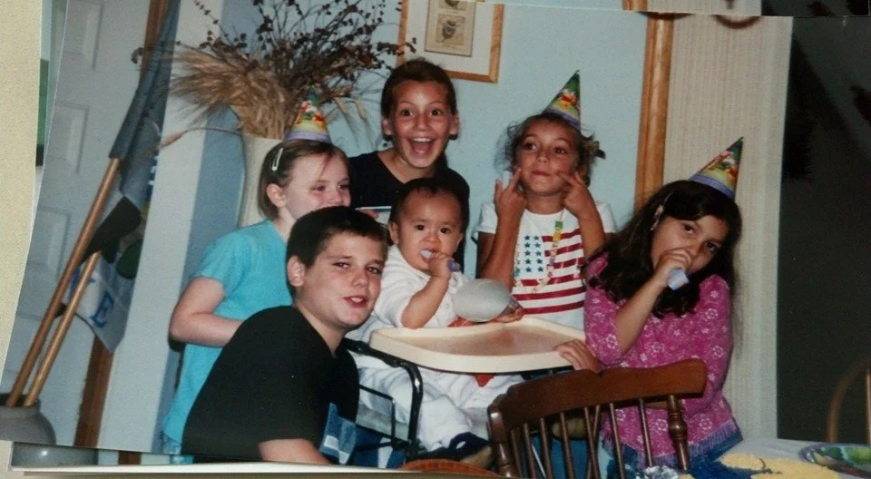 Group of six children celebrating birthday, wearing party hats, gathered around a table, one with a pacifier, smiling and posing for the photo.