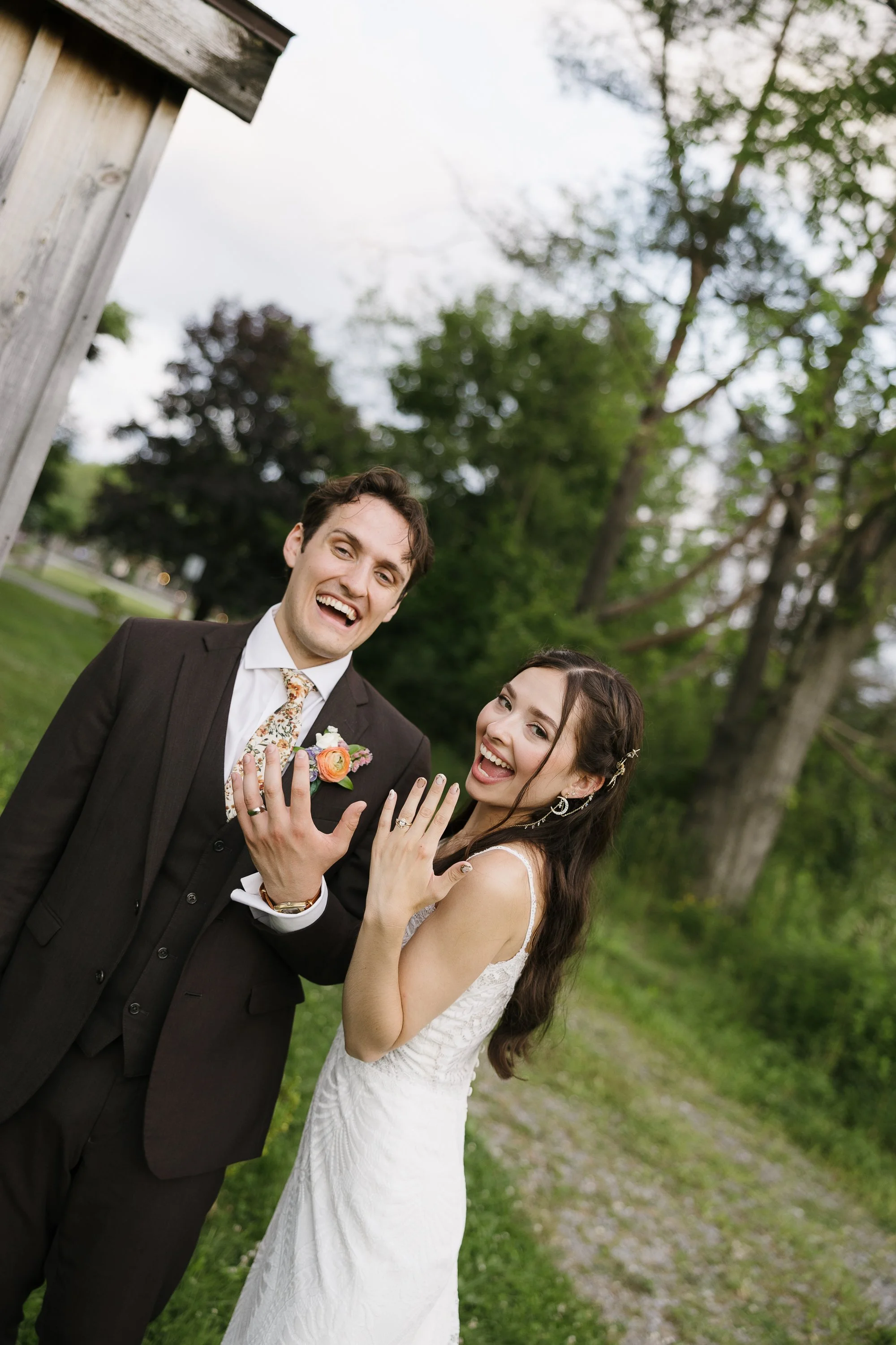 Happy couple in wedding attire showcasing wedding rings outdoors.