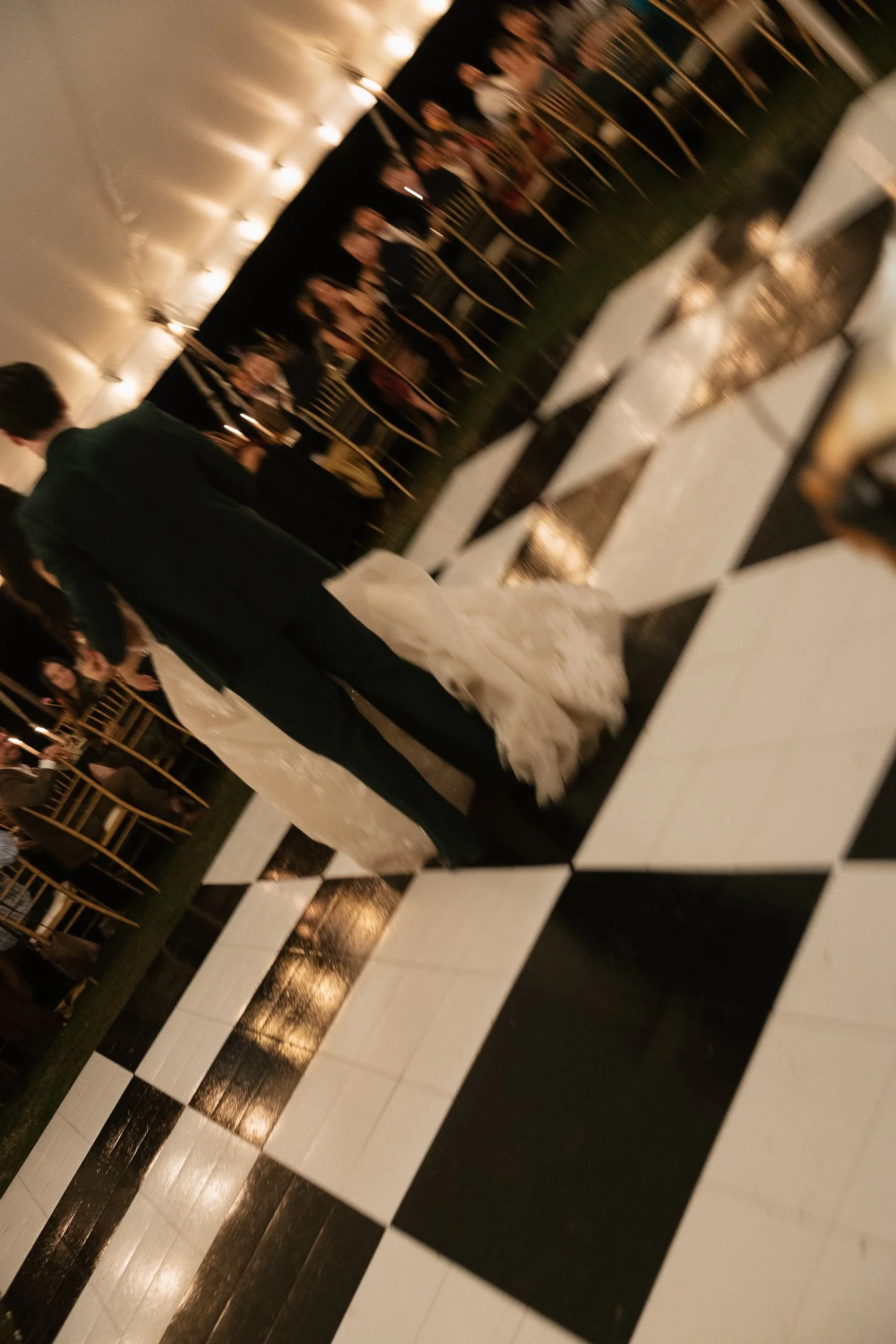 Couple dancing at a wedding reception with a checkered black and white floor and seated guests in the background.