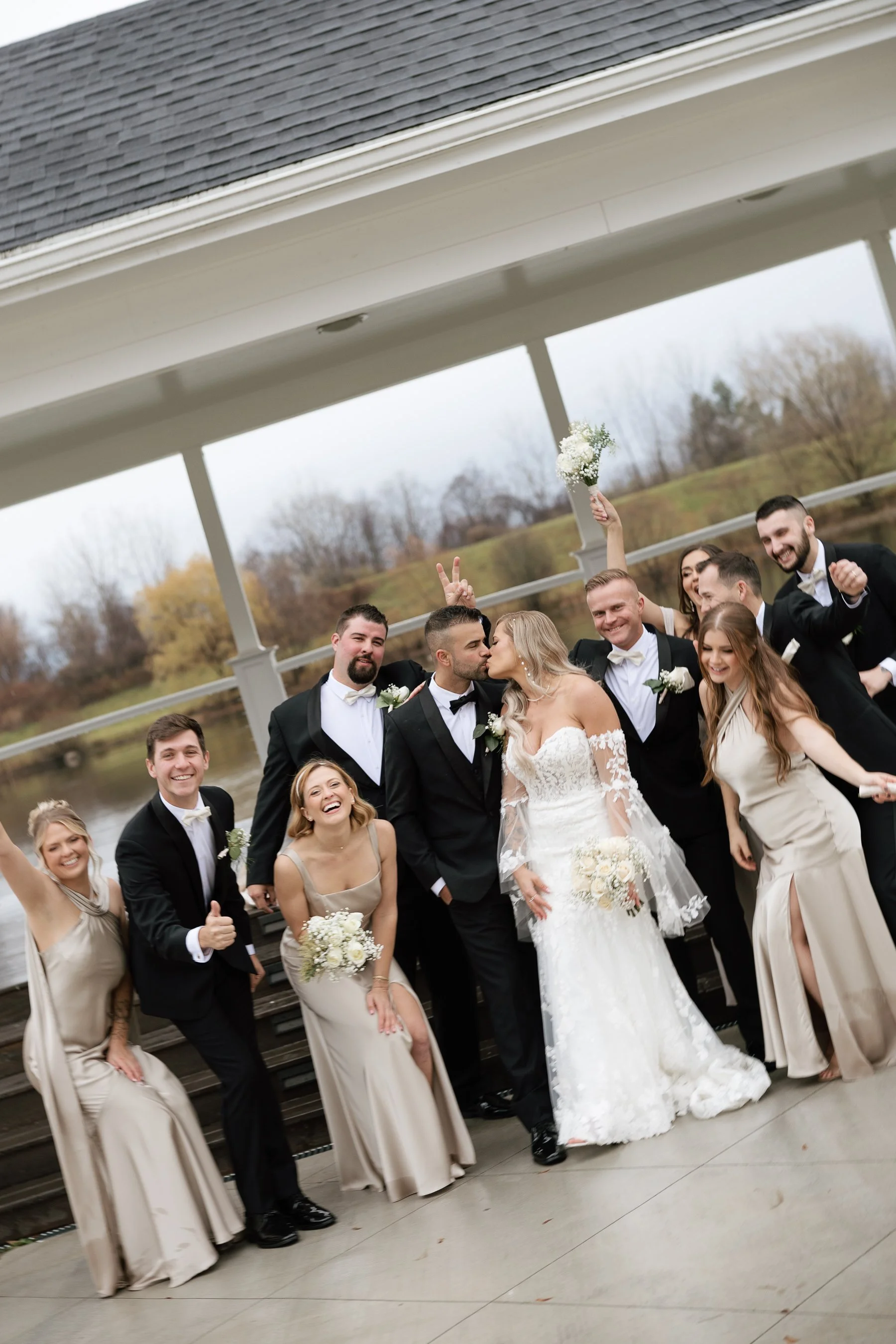 A group of wedding guests, including the bride and groom, celebrating outdoors near a lake with a bridge in the background. The bride is kissing the groom, and the guests are smiling and making playful gestures.