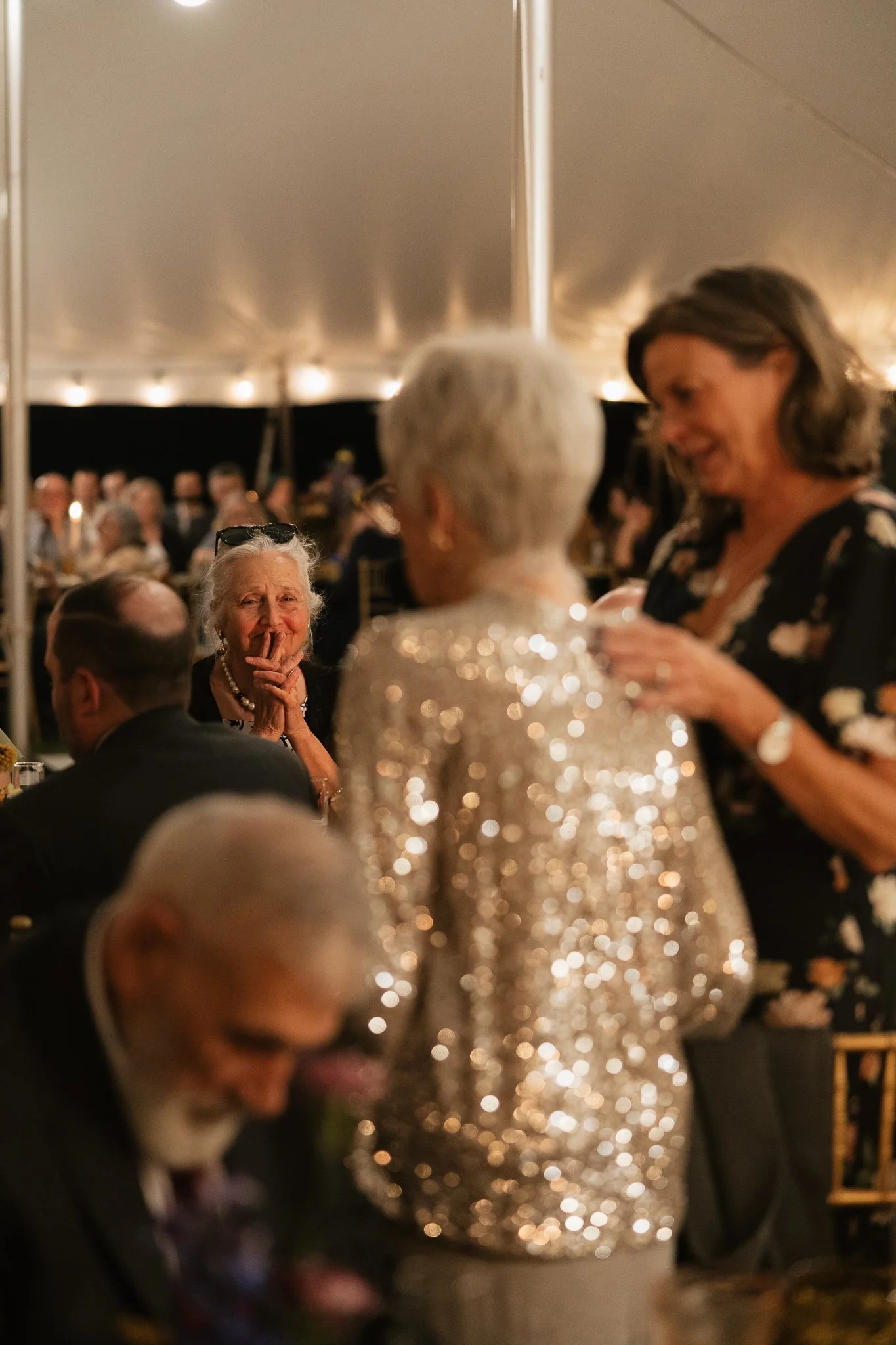 An elderly woman in a sparkling dress is talking to a woman in a floral dress at a formal gathering under a large tent, with other guests seated at tables in the background.