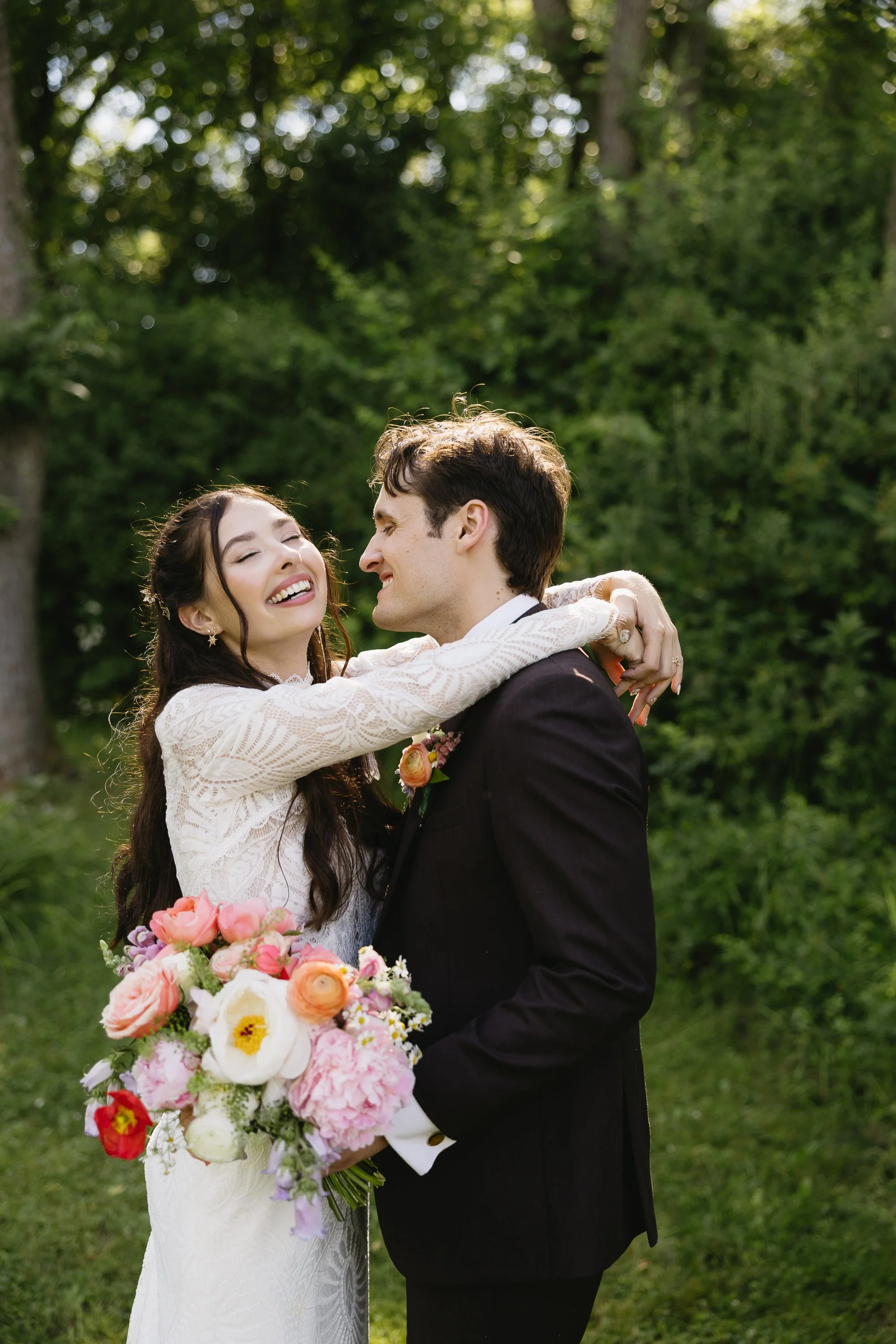 A bride and groom sharing a joyful moment outdoors, with the bride holding a colorful bouquet, in a lush green setting.