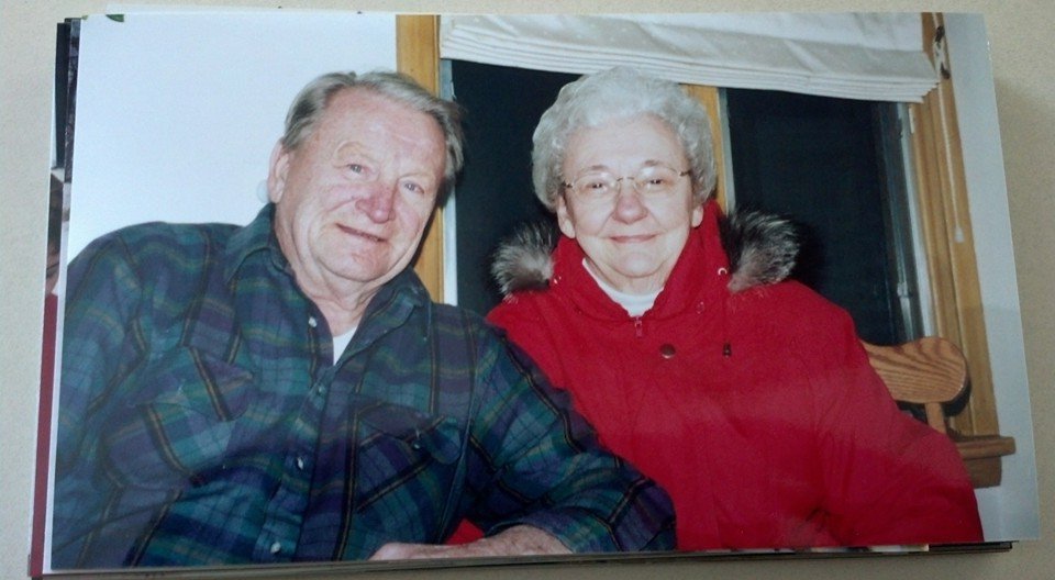 An elderly man and woman sitting close together, both smiling, in front of a window with a wooden frame. The man is wearing a plaid shirt, and the woman is wearing a red jacket with a fur-lined hood.