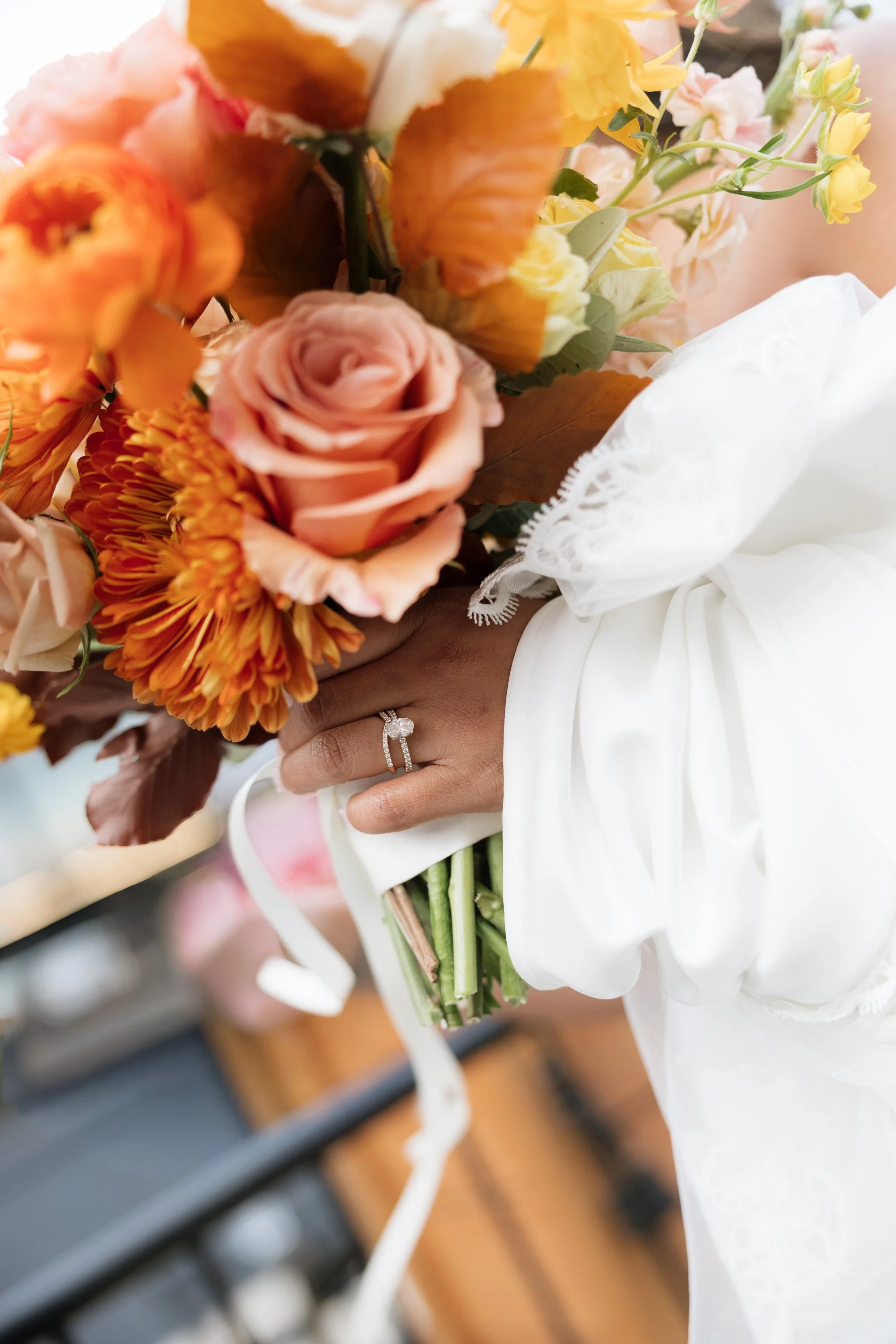 Close-up of a person holding a large bouquet of pink, orange, and yellow flowers, wearing a white lace dress and a wedding ring.