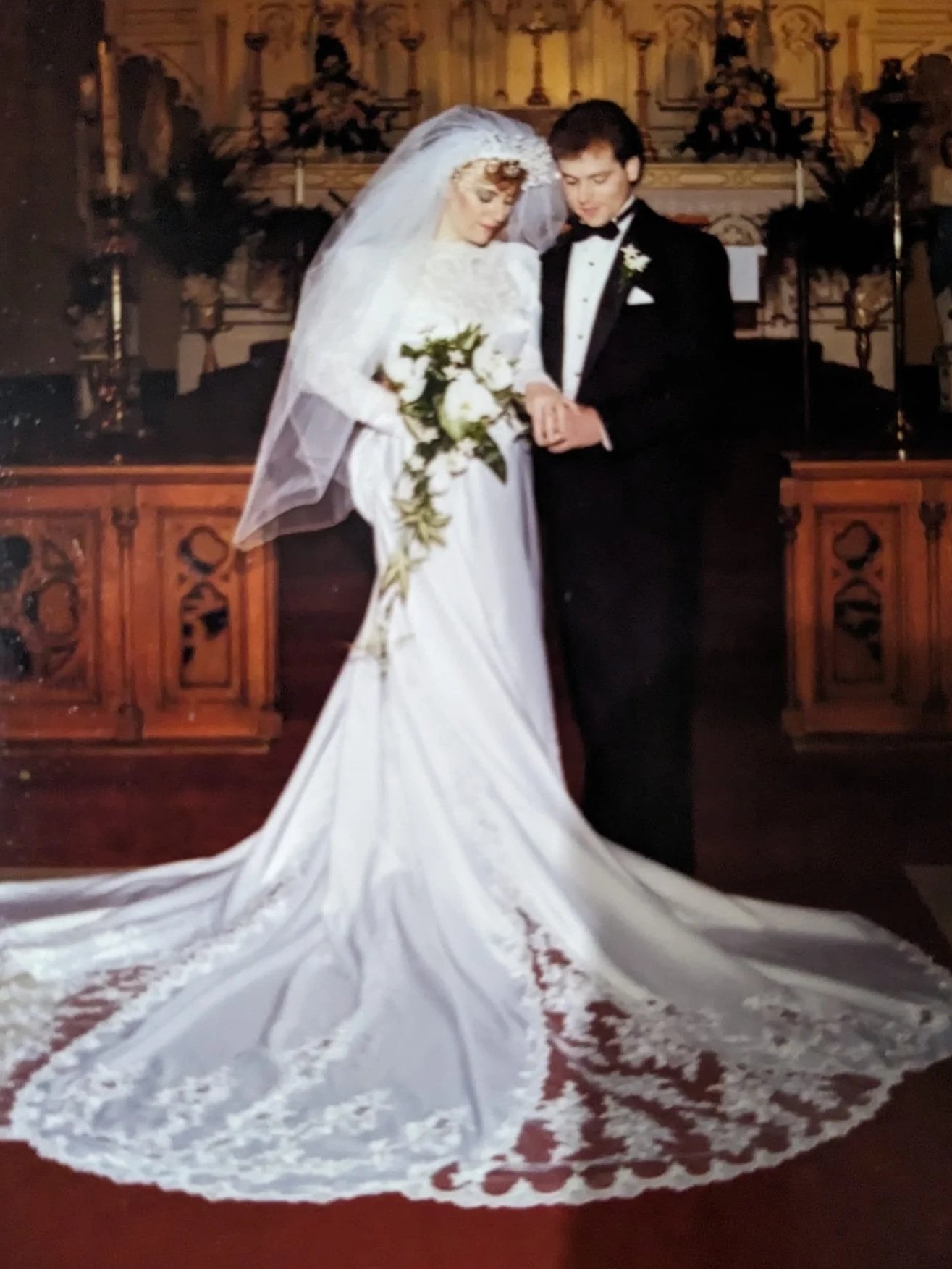 A bride and groom standing together in a church, holding hands, with the bride wearing a white wedding gown with a long train and veil, and the groom in a black tuxedo, during their wedding ceremony.