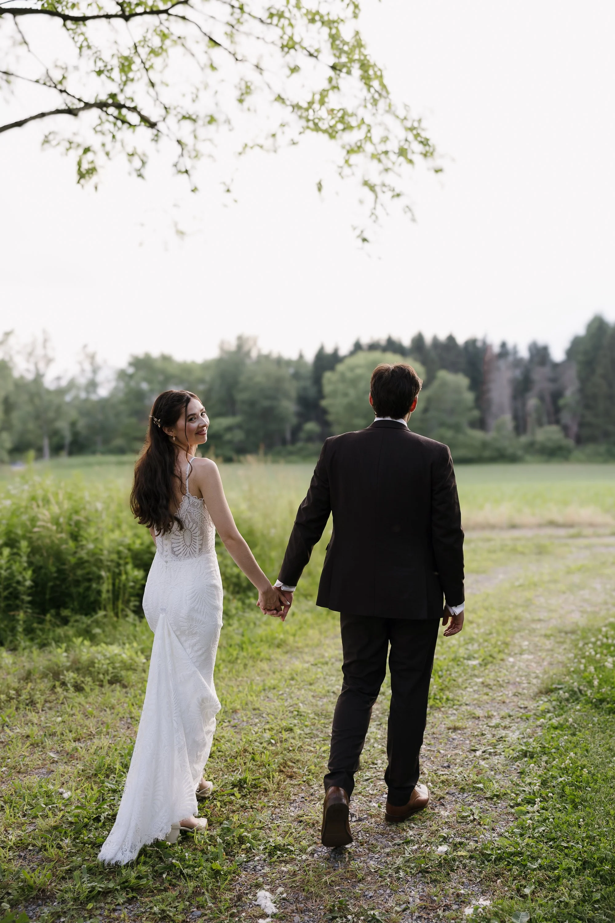 A bride and groom walking hand in hand through a park or field, with the bride looking back and smiling.
