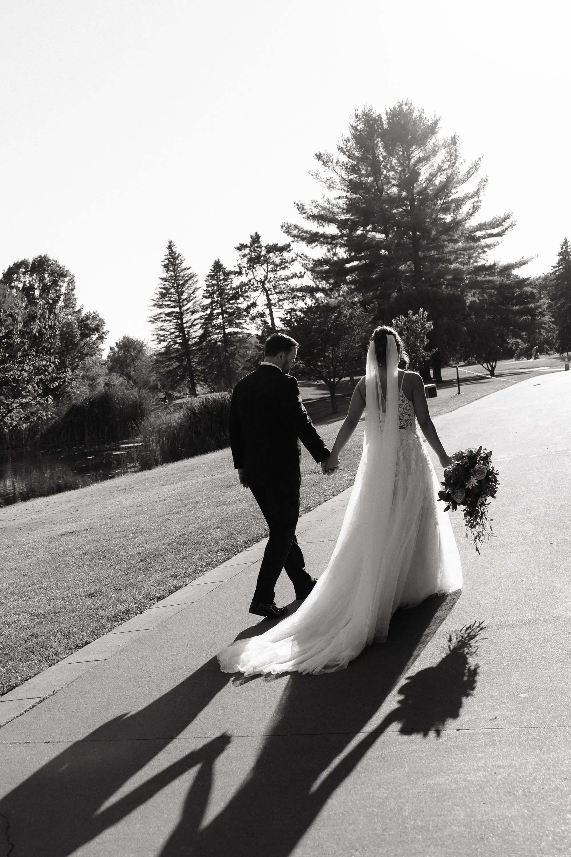 Black and white photo of a bride and groom walking hand in hand outdoors during a sunny day, with their shadows cast on the pavement, surrounded by trees and a park.