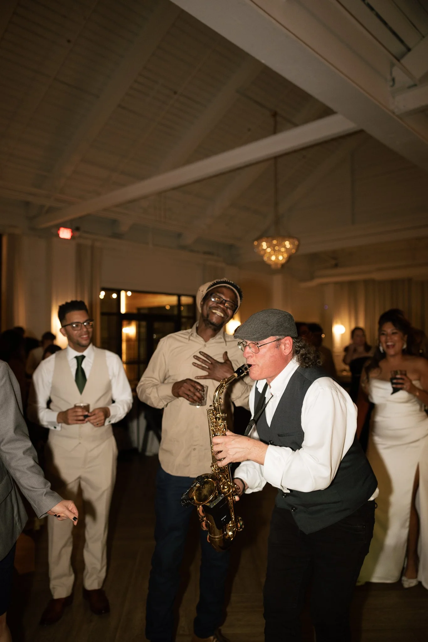 People at a celebration dancing and listening to a saxophone performance in an indoor venue with warm lighting and a chandelier.