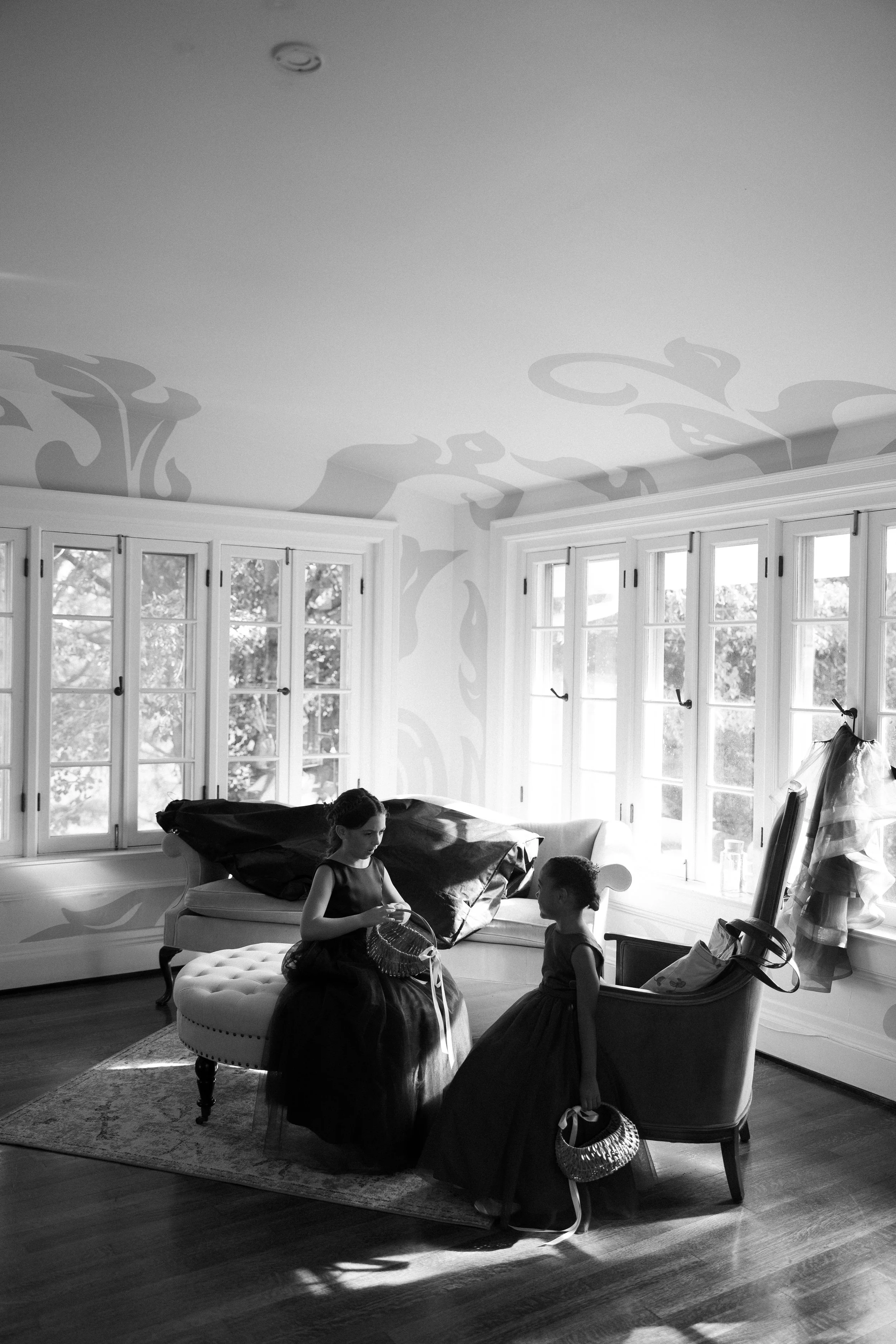 Two young girls in elegant dresses sitting in a sunlit room with large windows, talking and holding small handbags, with a couch and a wooden floor.