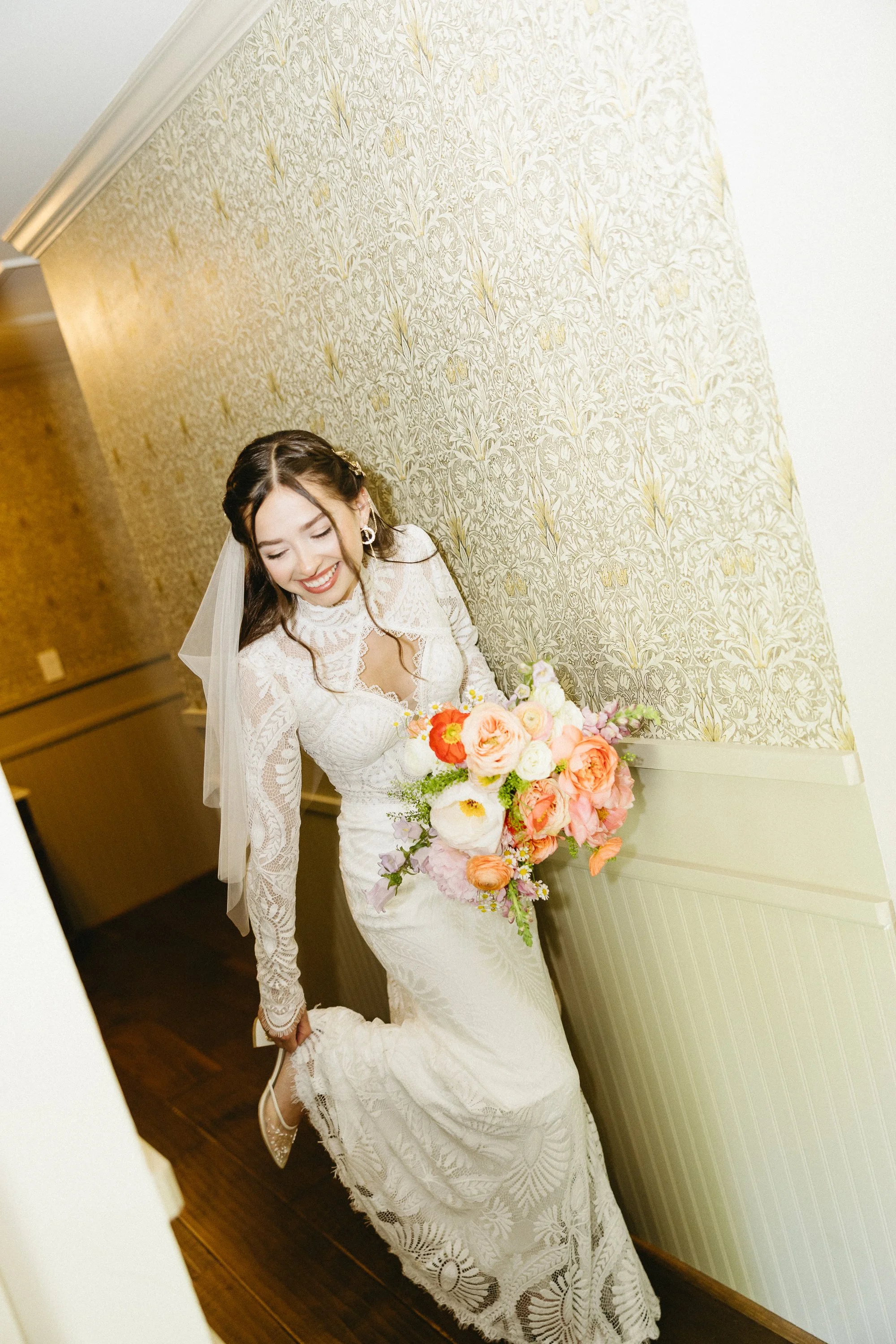 Bridal portrait of a smiling bride in a white lace wedding dress holding a bouquet of pink, white, and orange flowers, standing against a floral wallpaper wall.