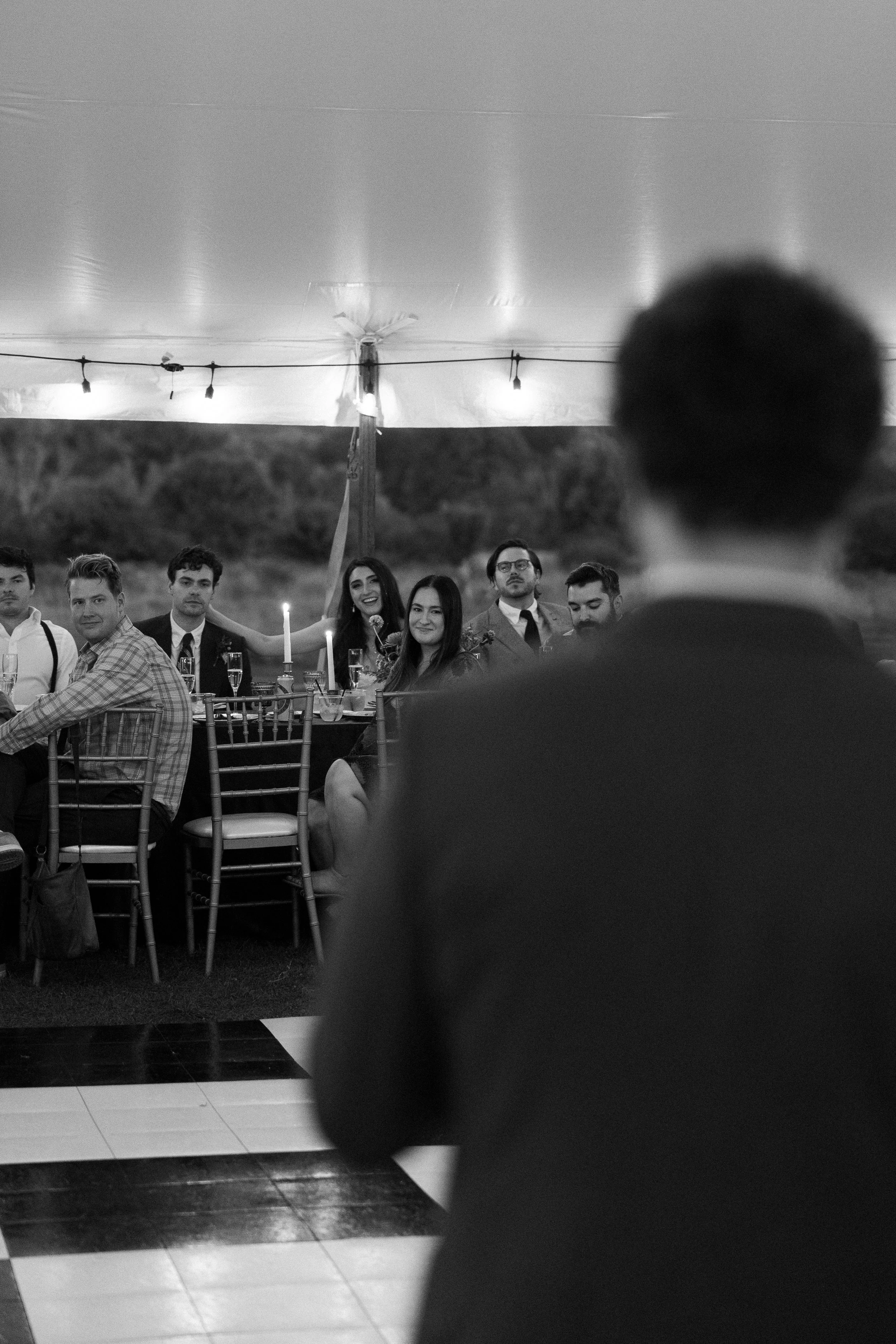 People seated at a table under a canopy, with a man in the foreground speaking to the group, at an outdoor event during evening.