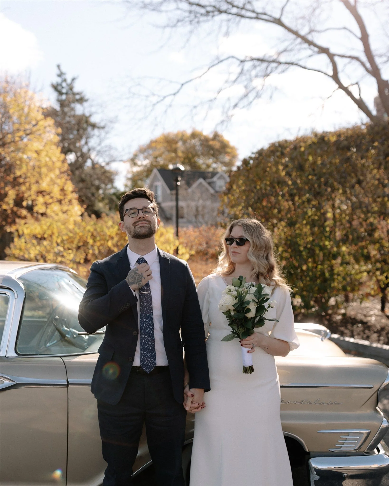 A man in a suit holding a woman's hand, standing next to a vintage car outdoors, with trees and a house in the background during fall.