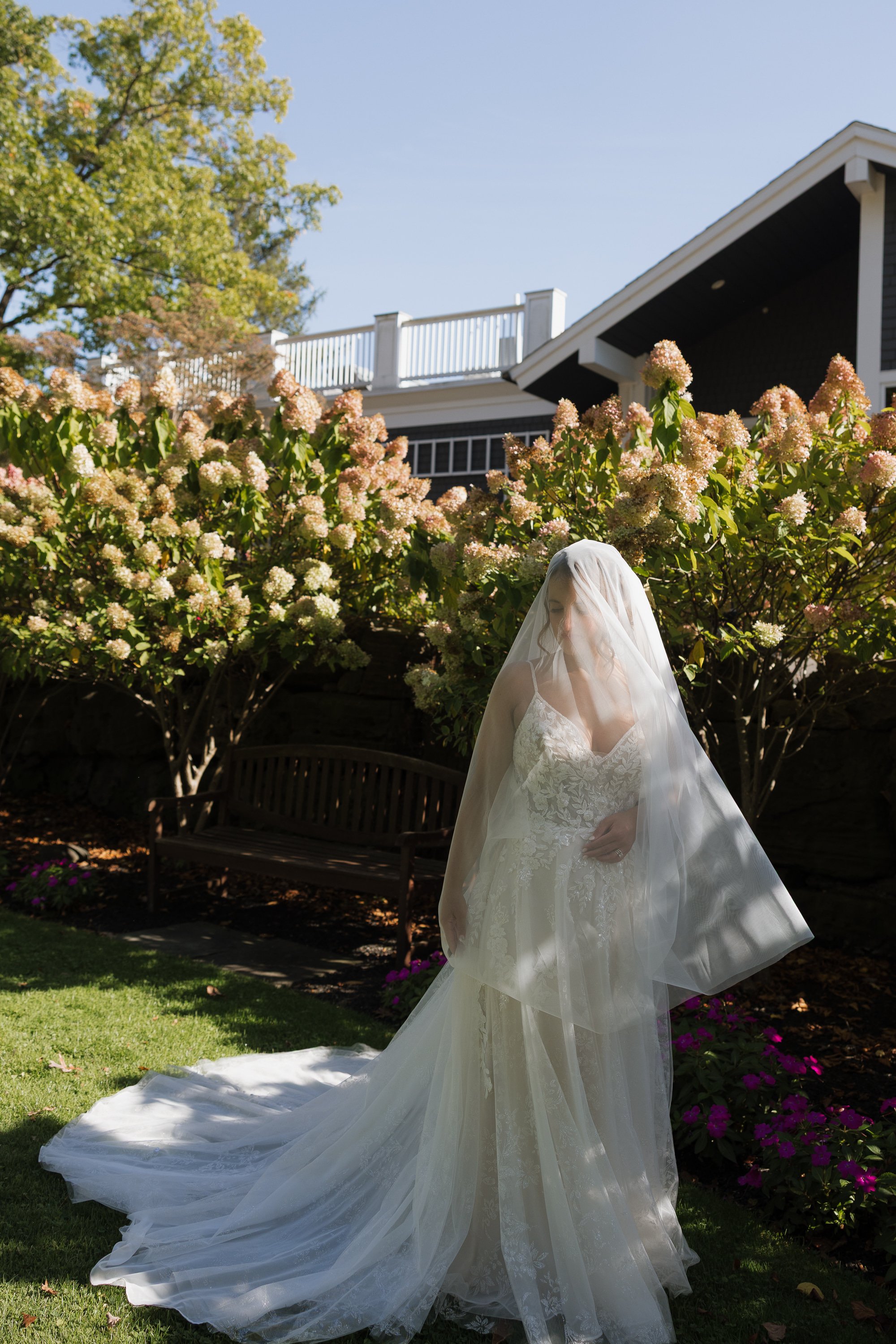 A bride in a white wedding dress with a veil, standing in a garden with blooming pink and white flowers, a wooden bench, and a house with a white fence in the background.