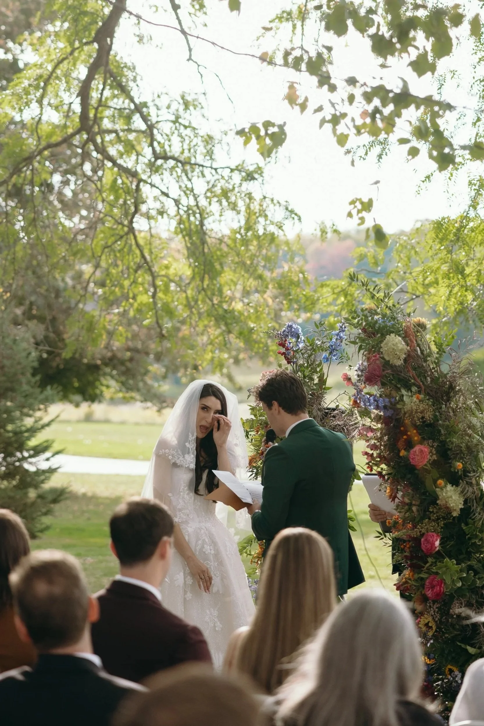 A bride wiping tears at an outdoor wedding ceremony, standing next to the groom with a colorful floral arrangement behind them, while guests watch.