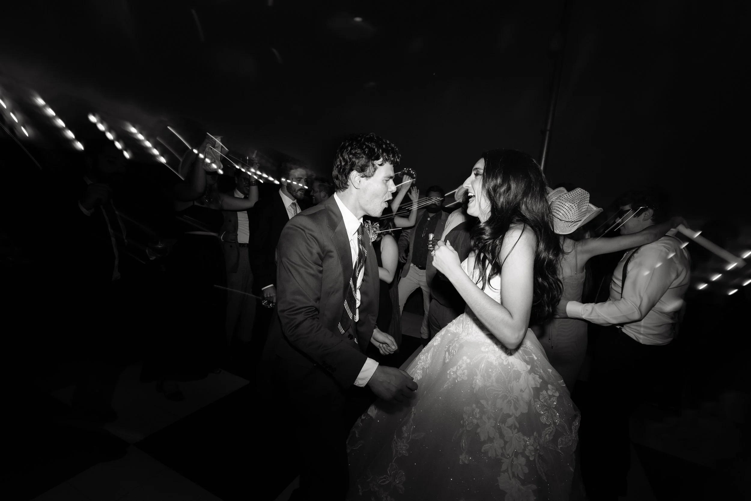 Black and white photo of a bride and groom dancing closely at a wedding reception, surrounded by guests.
