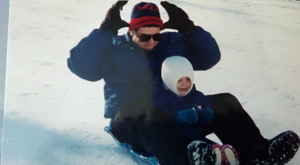 An adult man and a young girl sitting on a sled on snow, wearing winter clothing and smiling.