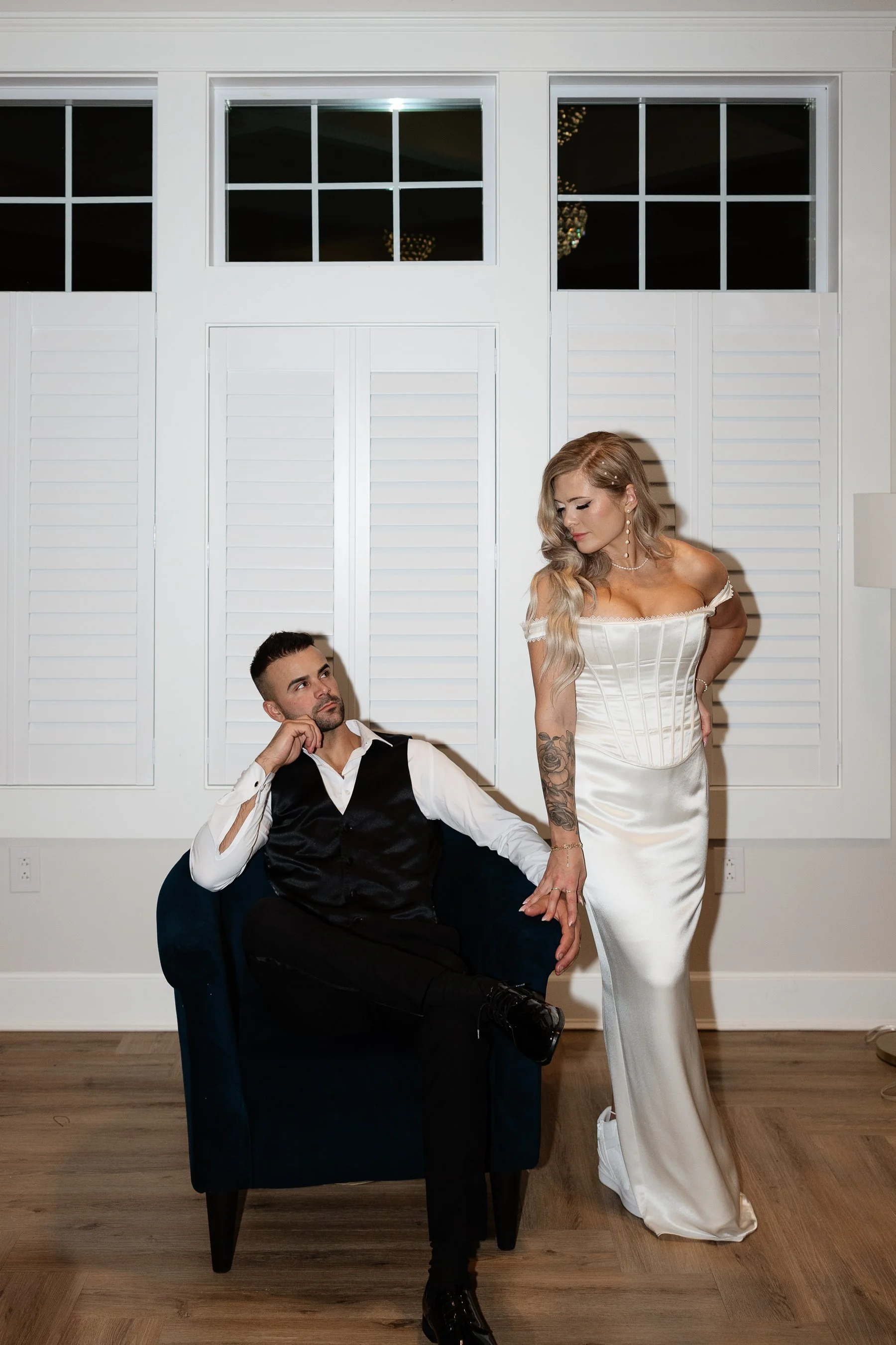 A woman in a white wedding gown standing next to a seated man in a white shirt and black vest, in a room with white walls and windows with shutters.