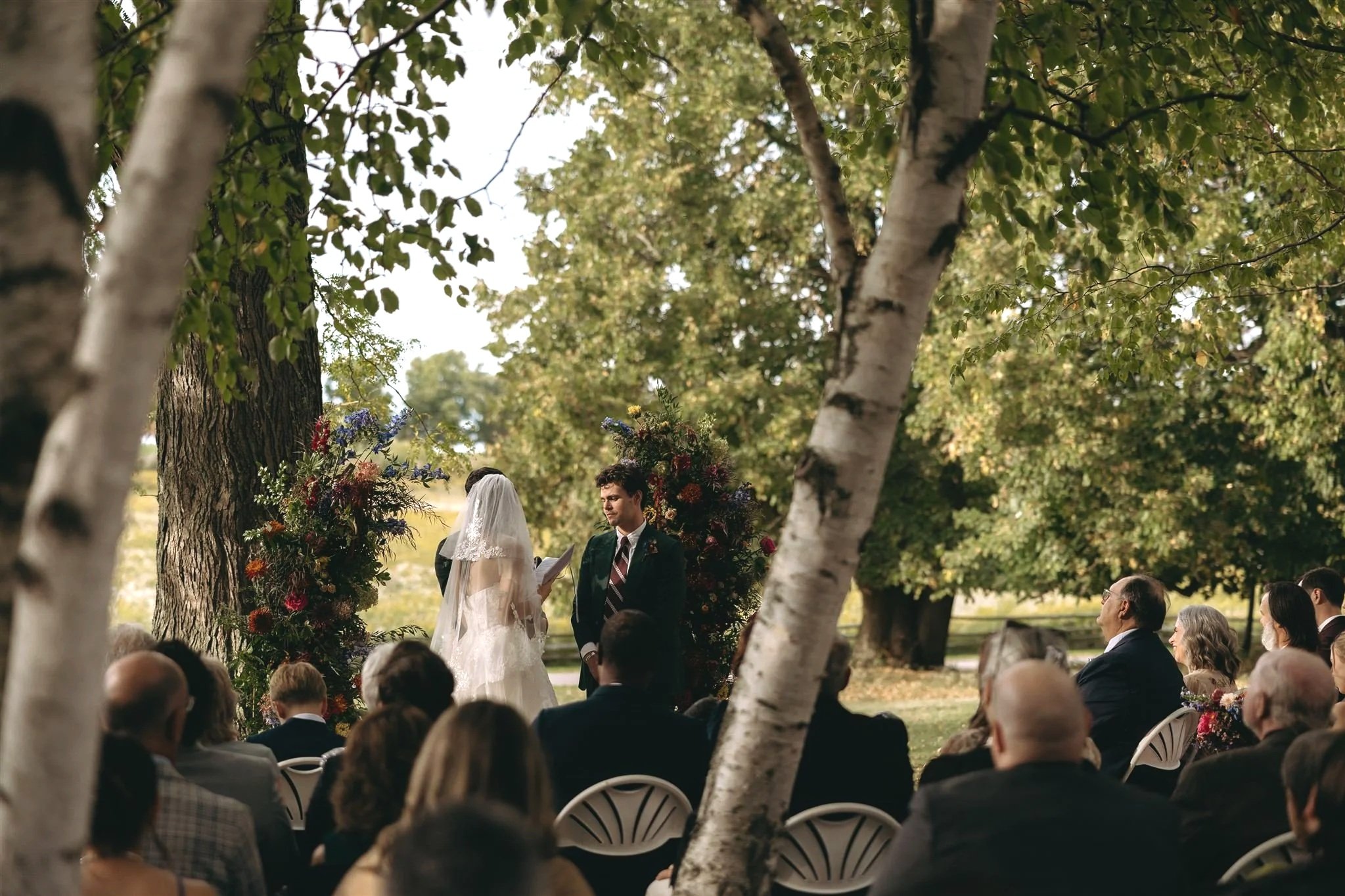 A wedding ceremony taking place outdoors under trees with a bride and groom facing each other, surrounded by guests seated in white chairs, and floral arrangements in the background.