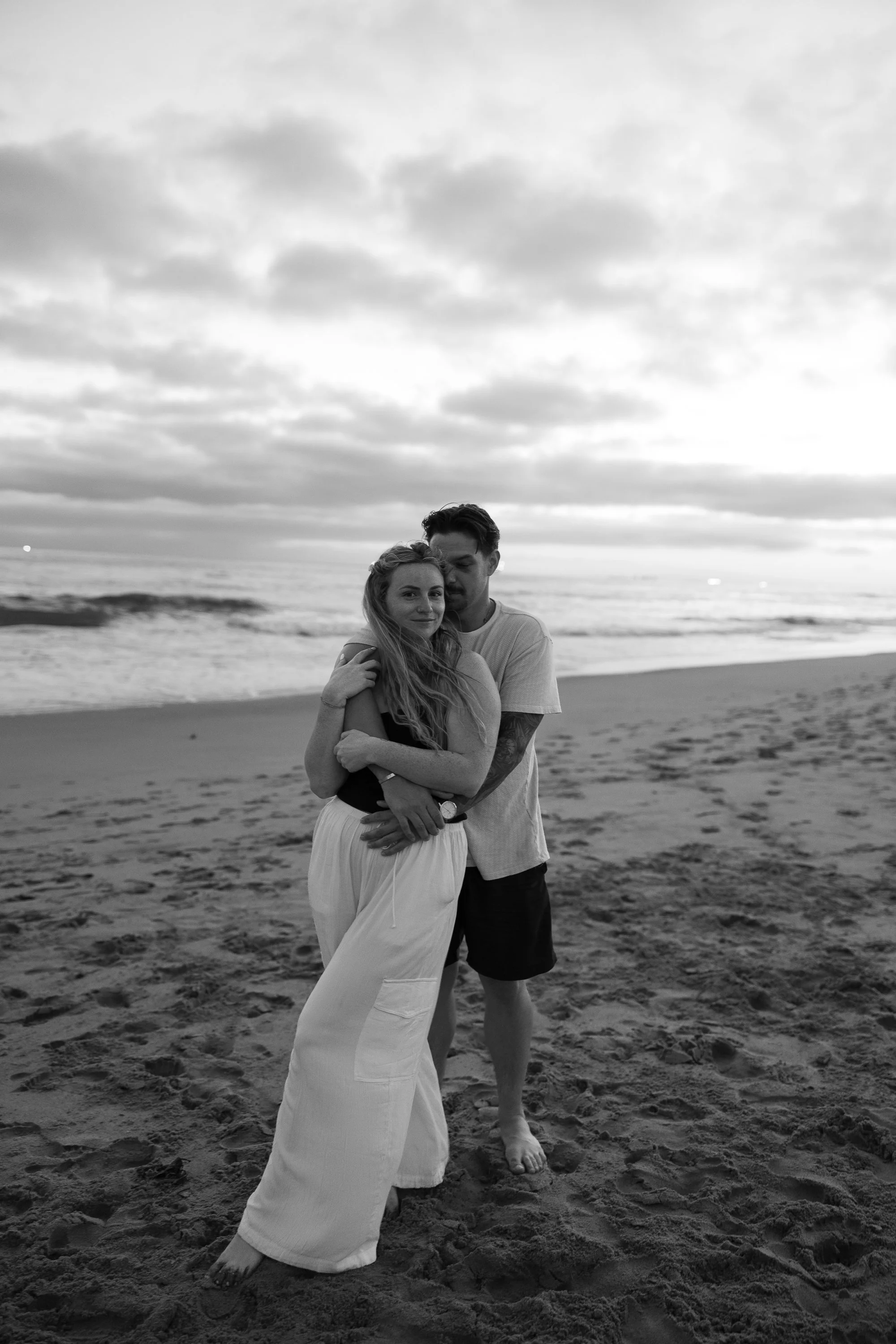 A black and white photo of a man and woman embracing on a beach, with ocean waves and cloudy sky in the background.
