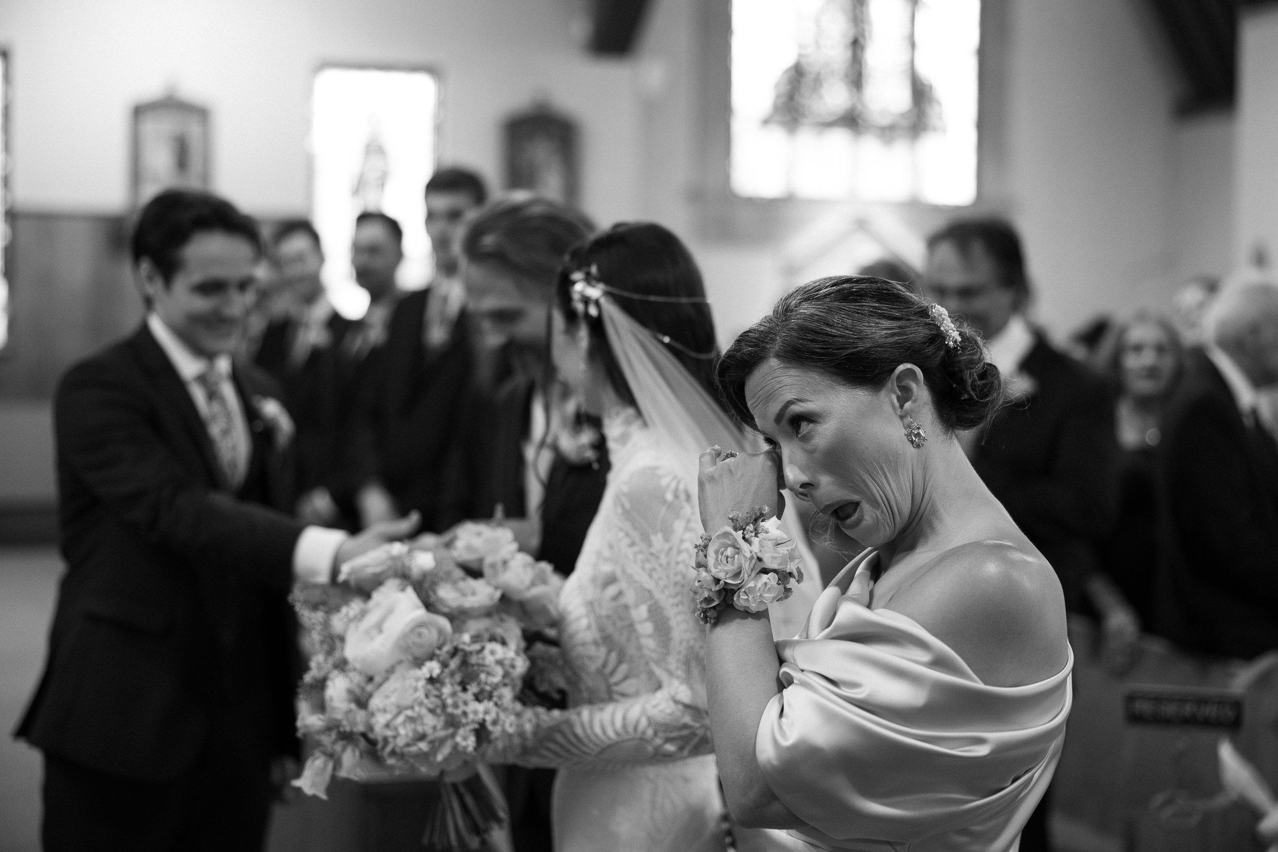 A woman in wedding attire appears emotional during a ceremony, with a bridesmaid holding flowers beside her, and groom and guests in the background inside a church.