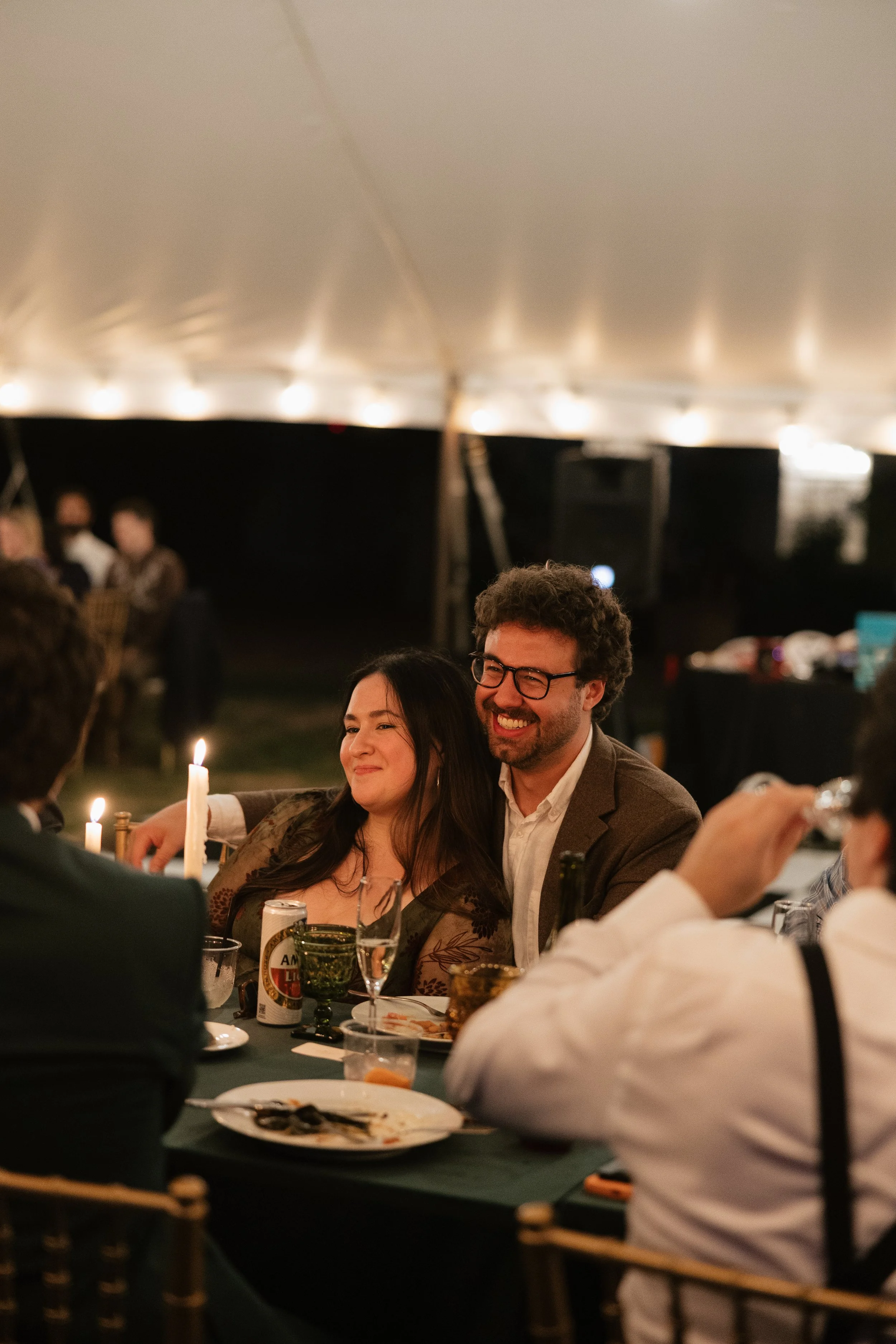 People at a dinner party sitting under a tent, smiling and enjoying drinks, with candles on the table.
