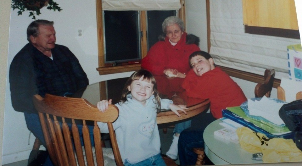 Family gathered around a wooden dining table, smiling, with a young girl in the foreground and an elderly woman and two boys in the background.