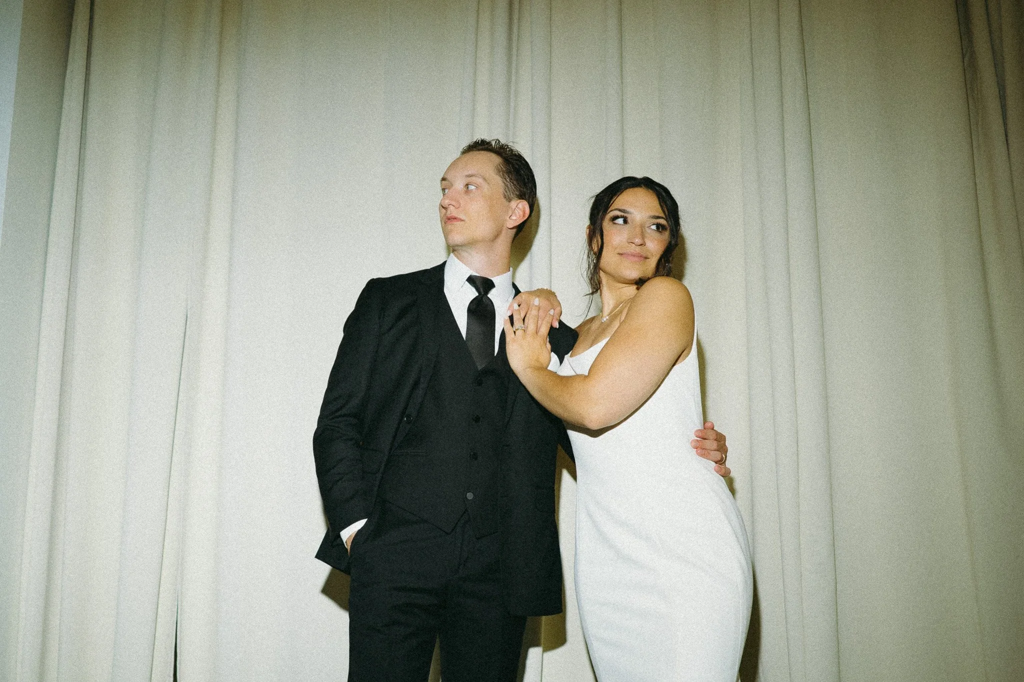 A couple dressed in formal attire, the man in a black suit and tie, the woman in a white dress, standing close together against a plain white wall.