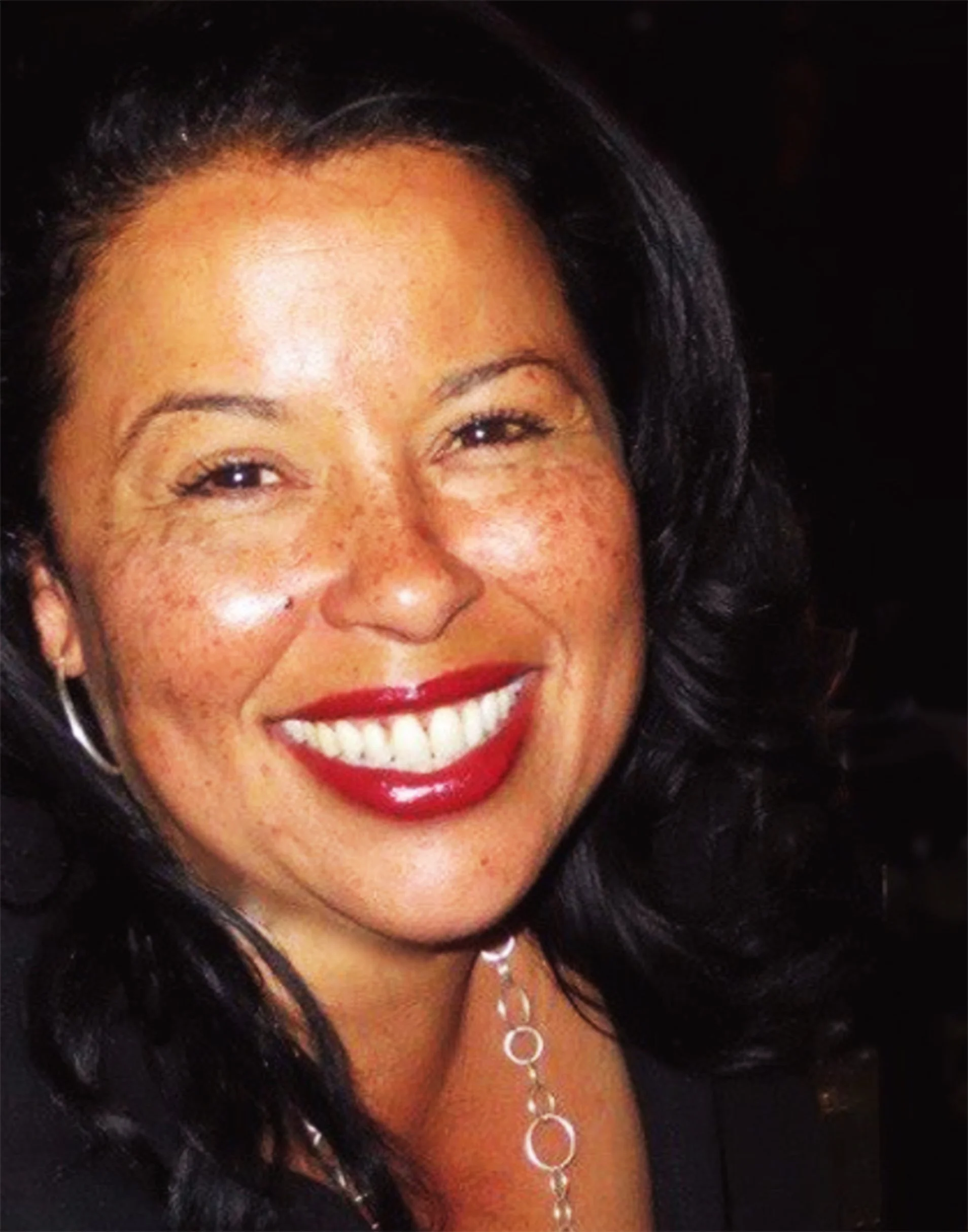 A woman smiling with dark hair and red lipstick, wearing earrings and a necklace, against a dark background.