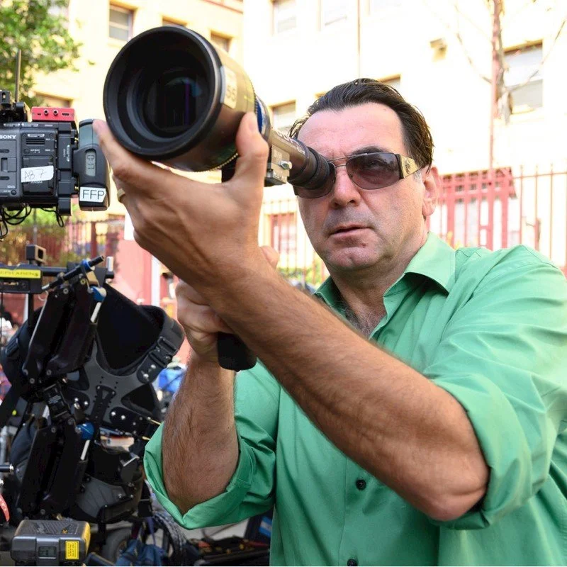 A man wearing sunglasses and a green shirt using a professional video camera with a large lens outdoors.