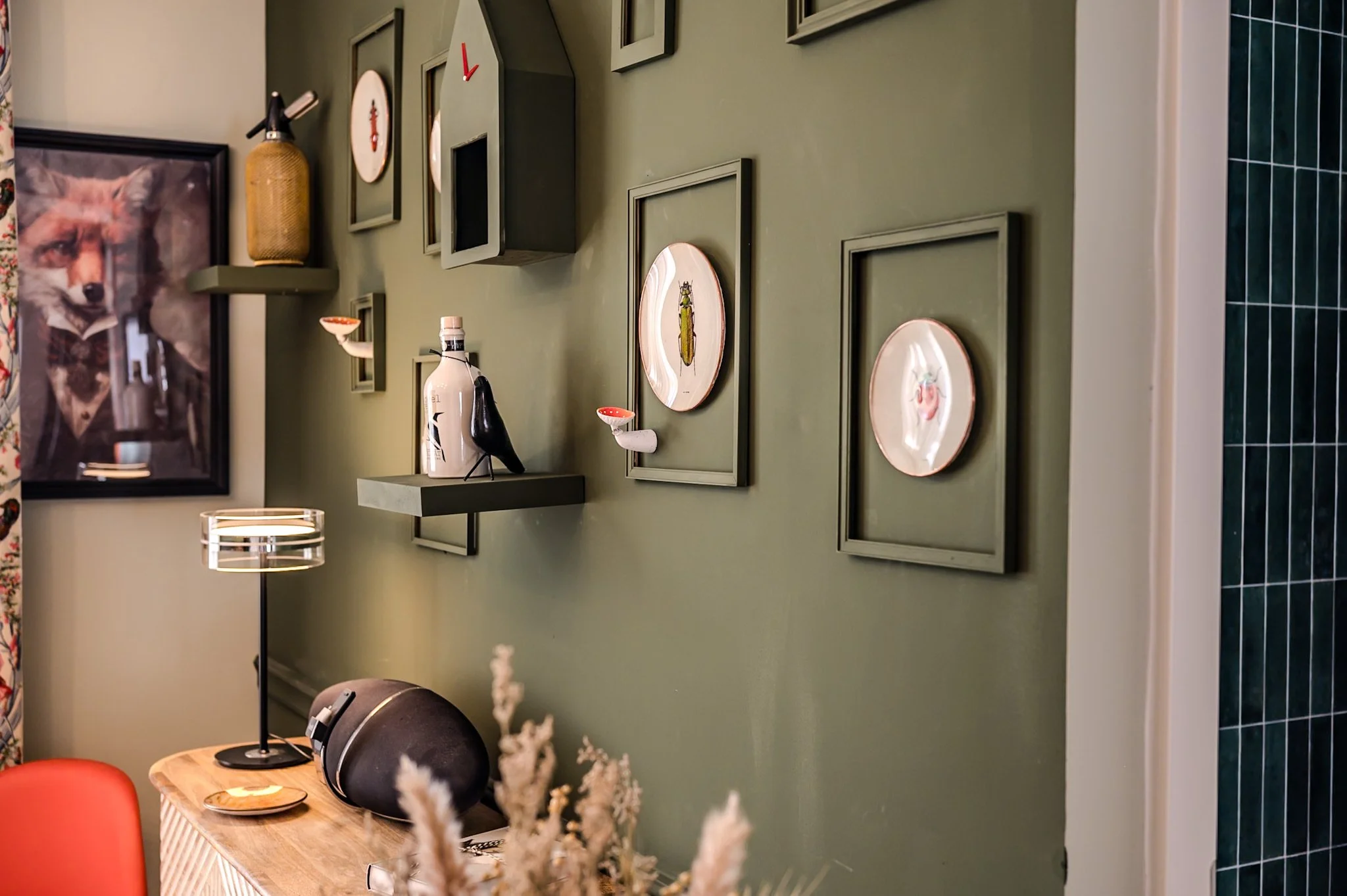 Ein Wohnzimmer mit grün gestrachter Wand, dekorativen Bilderrahmen, einer Flasche und Flaschenschirm auf einer schwarzen Ablage, einem Tischlampe, einem Sessel und einem Bild eines Fuchses an der Wand.