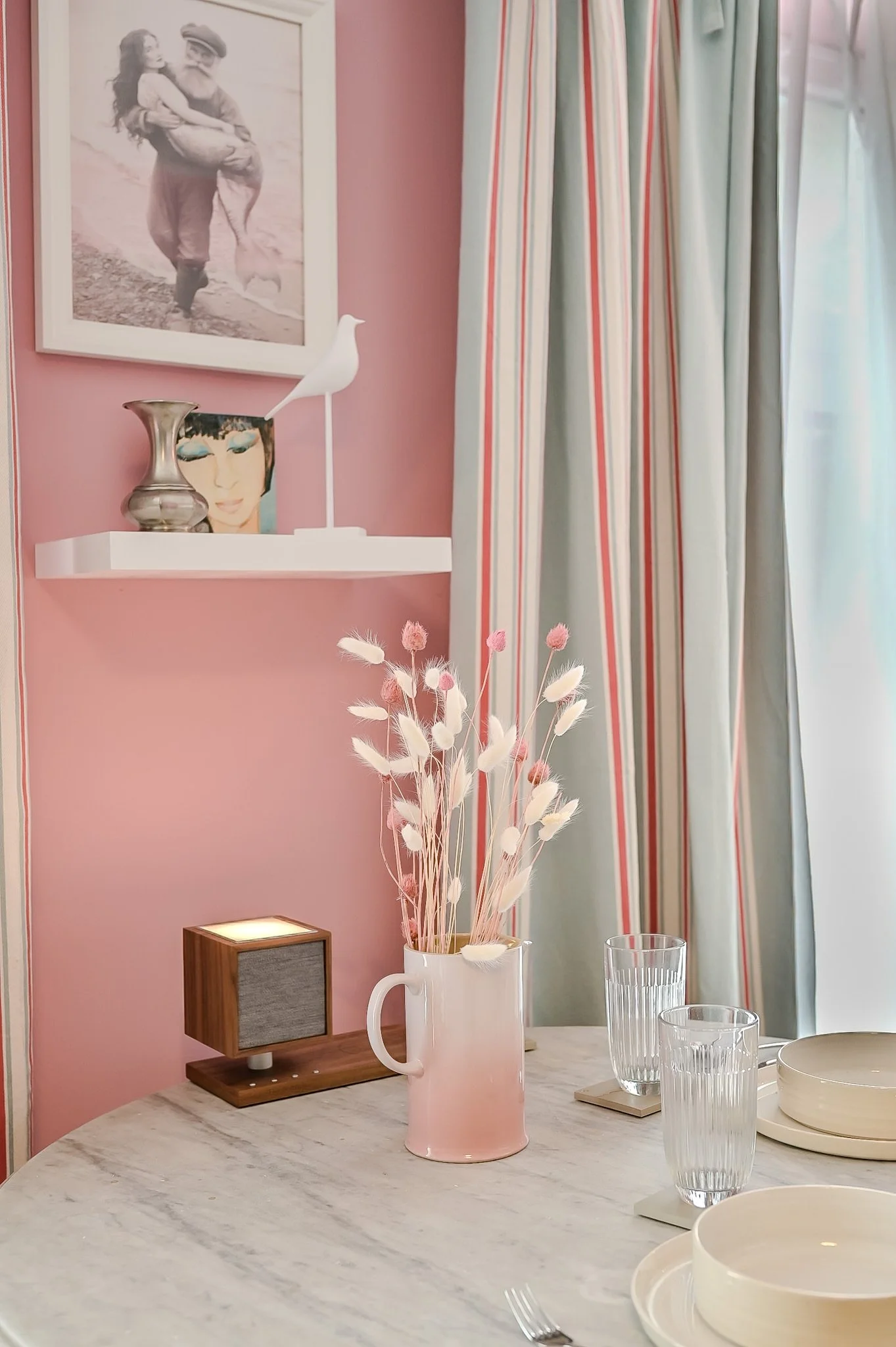 Ein Esstisch mit weißen Tellern, Gläsern und einer rosa Vase mit getrockneten Blumen, vor einem Fenster mit gestreiften Vorhängen, an einer pinken Wand mit einer weißen Galerie-Schiene mit Kunstwerken und Dekorationen.