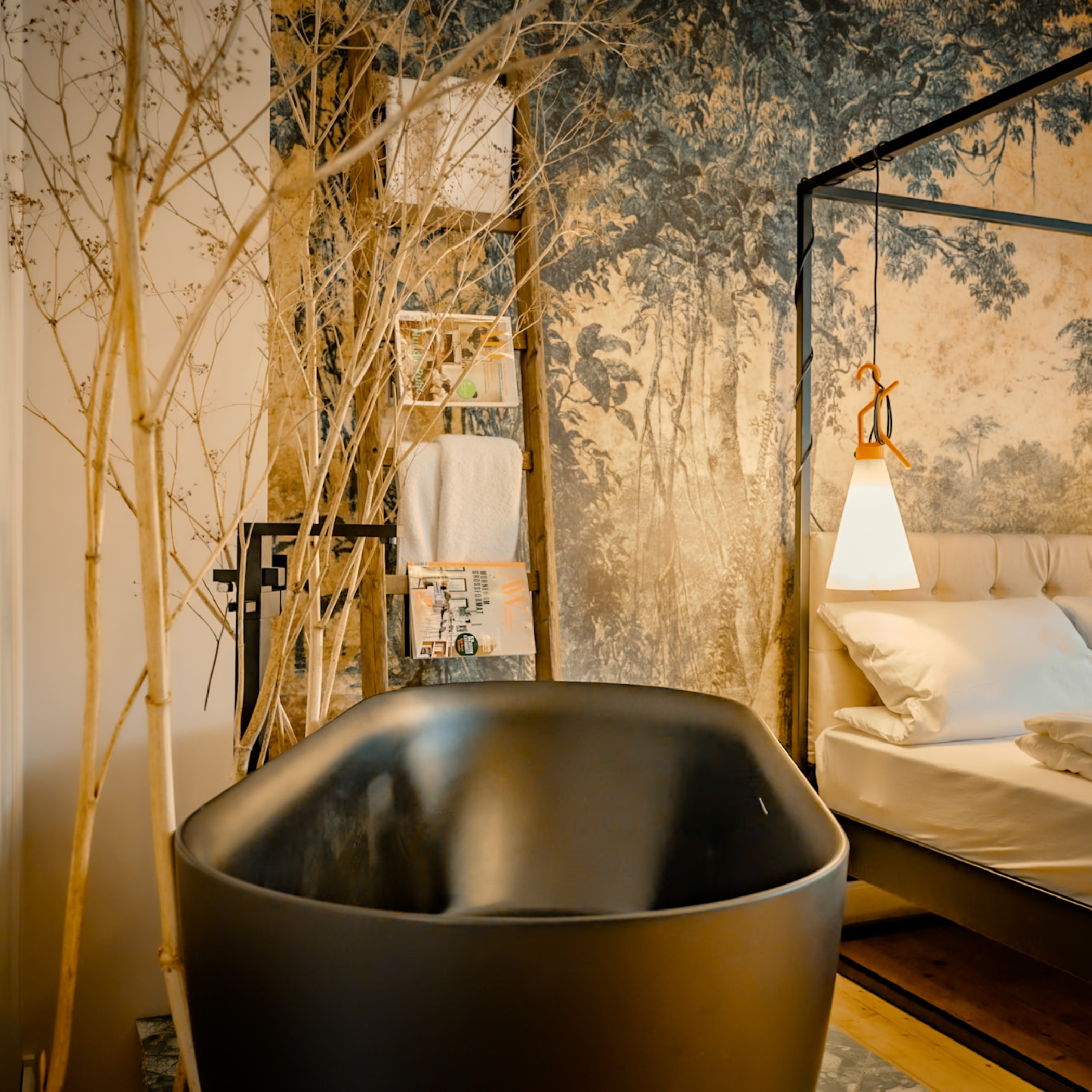 Ein modernes Schlafzimmer mit einer Badewanne aus schwarzem Material, einem aufblasbaren Bett mit weißen Bettlaken, einer Leseleuchte, Wanddekoration mit Waldmotiv und einer kleinen Holzregal mit Zeitschriften, Handtüchern und Pflanzen.