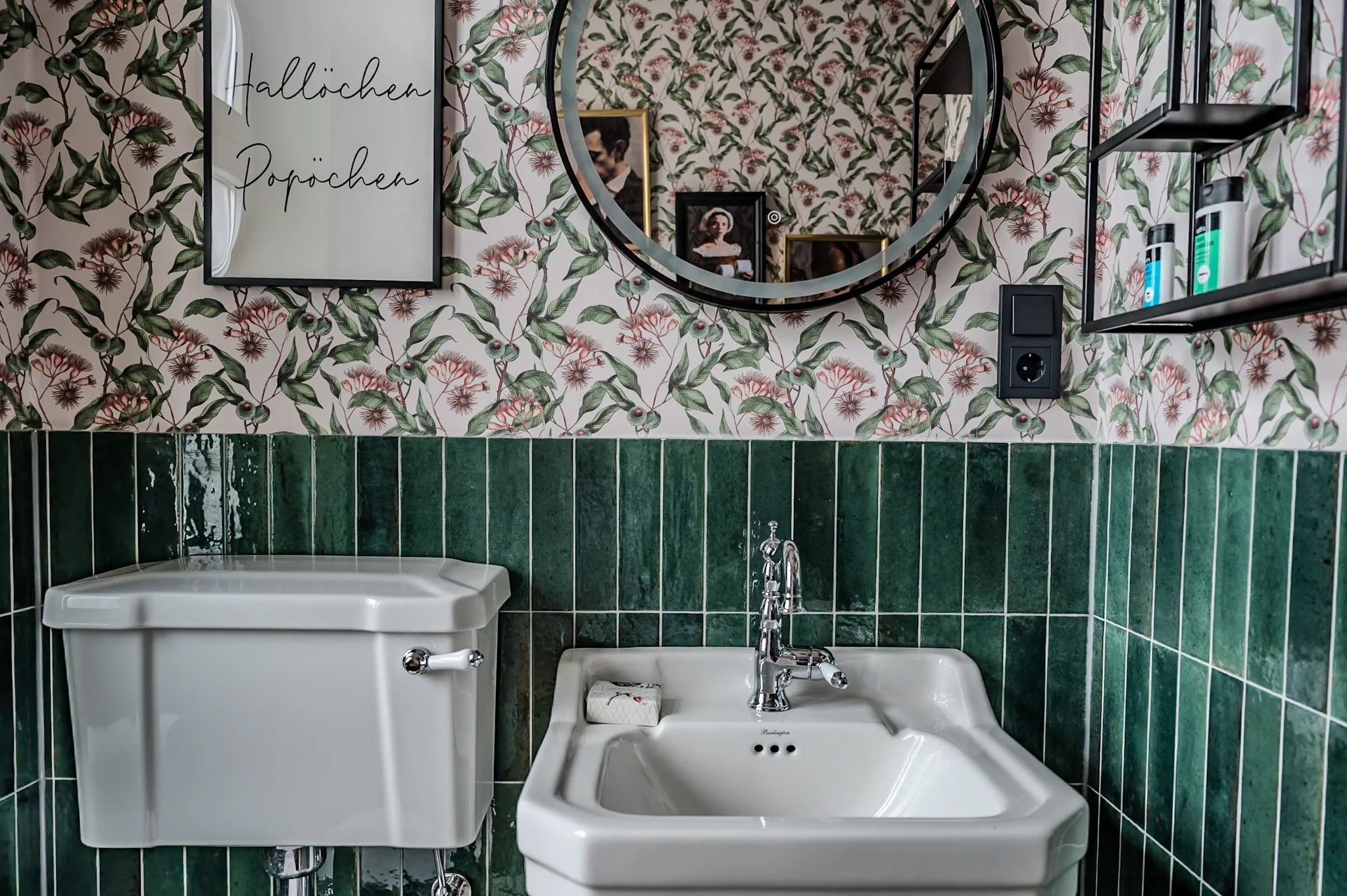 Ein Badezimmer mit grünen Fliesen, einem weißen Waschbecken mit Wasserhahn, einem uralten Spülkasten, einem runden Spiegel, Wandbildern und offenen schwarzen Regalen mit Pflegeprodukten. Die Wand ist mit einer floral- und blättrigen Tapete dekoriert,