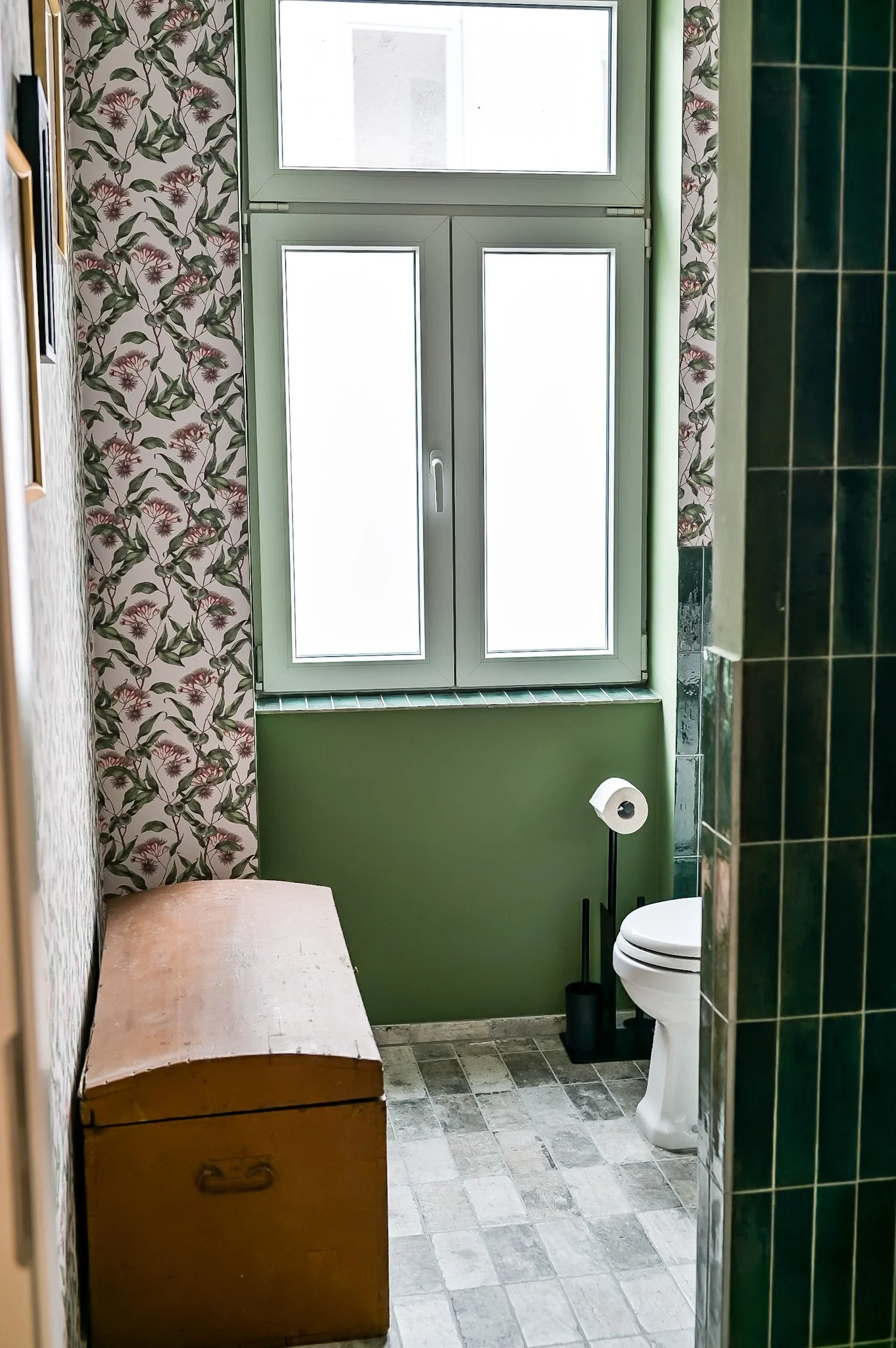 Ein Badezimmer mit grünen Wänden, einem Fenster, einem Toilettenpapierhalter, einem alten Holzkoffer und grünen Fliesen am rechten Wande. Der Blick ist durch die Tür ins Badezimmer gerichtet.