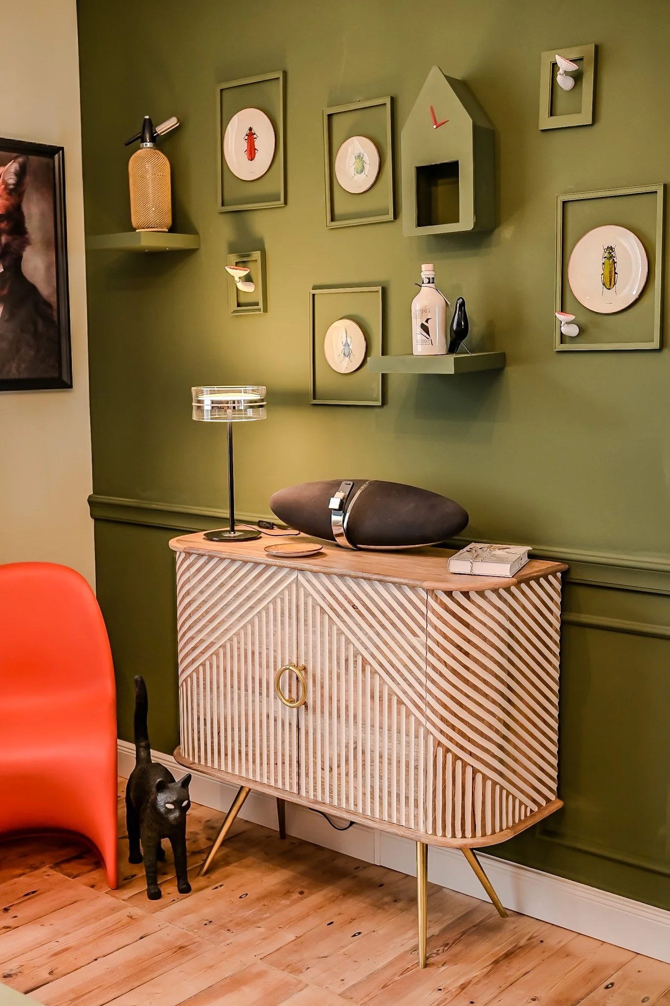 Ein stilvolles Wohnzimmer mit einer grünen Wand, dekoriert mit Bildern und Deko-Objekten, einem Holzsideboard mit goldenen Beinen, einem schwarzen Lautsprecher und einem Tischlampenschirm. Ein schwarze Katzenfigur steht neben einem orangefarbenen Stu
