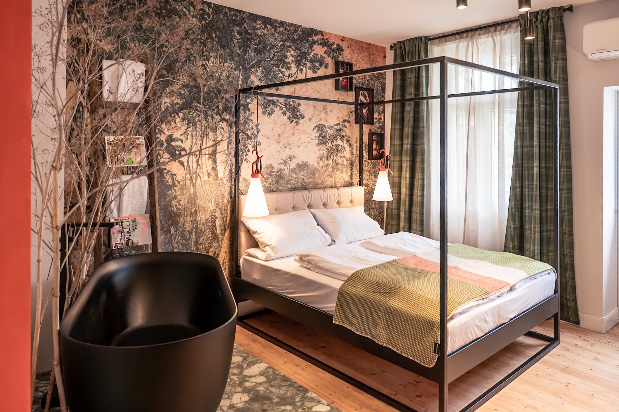 Schlafzimmer mit Himmelbett, Wandtapete mit Jungle-Motiv, schwarz-weißer Baumstamm und Bilder an der Wand, Fenster mit grünen Gardinen, schwarzer Wannenstuhl, weiße Bettwäsche und bunte Tagesdecke, helle Holzdielenboden.